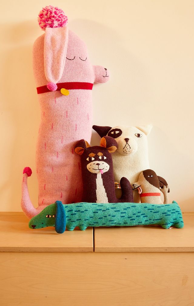 A collection of handmade plush toys and a decorative pillow arranged on a light wooden shelf against a warm beige wall. The collection includes a tall pink llama-shaped pillow with a pom-pom, a green crocodile-shaped pillow, and several small animal plushies including a white cat, a brown cat, and a poodle.