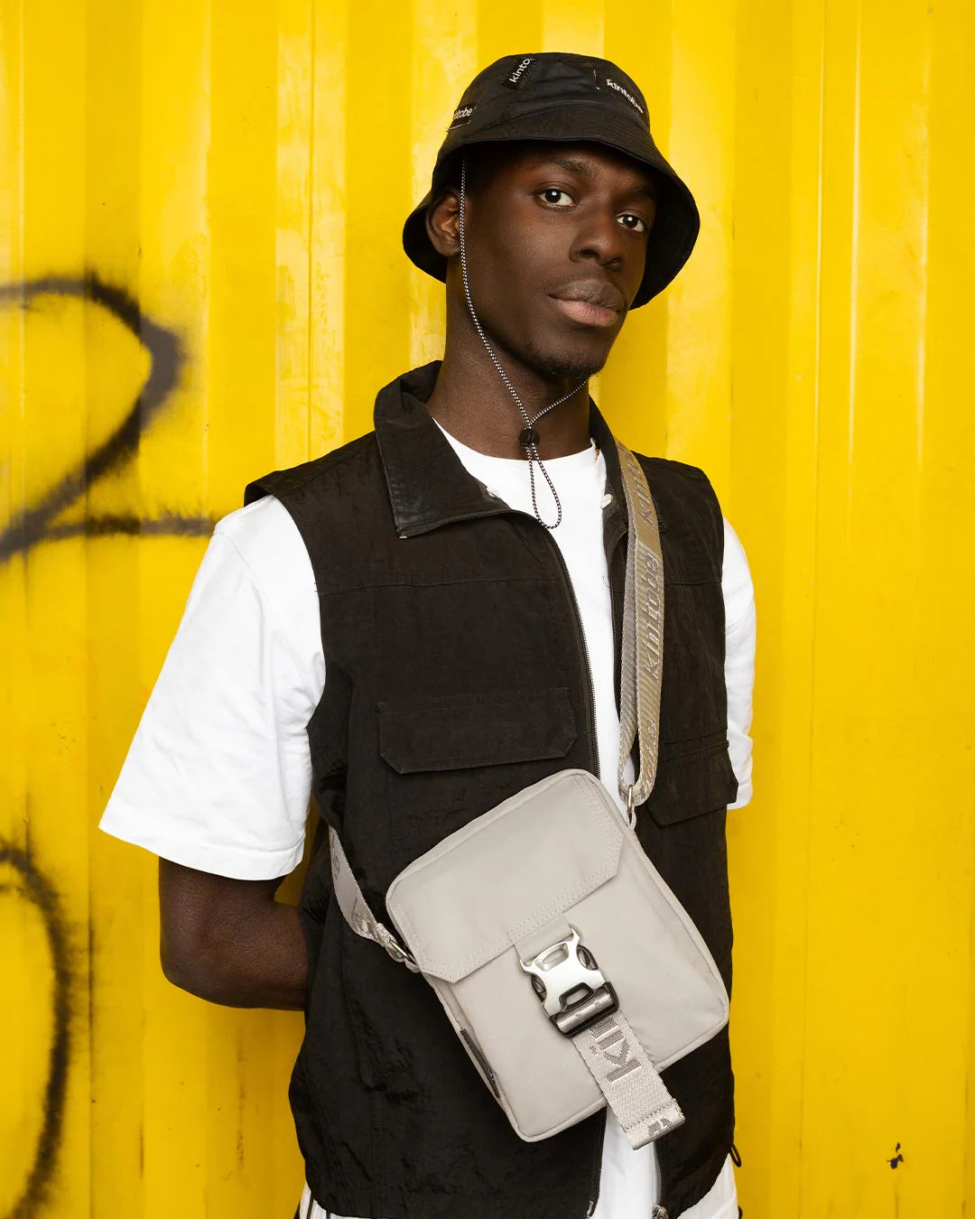 Light gray crossbody bag with silver buckle and strap, worn by a person in a black vest against a yellow background.