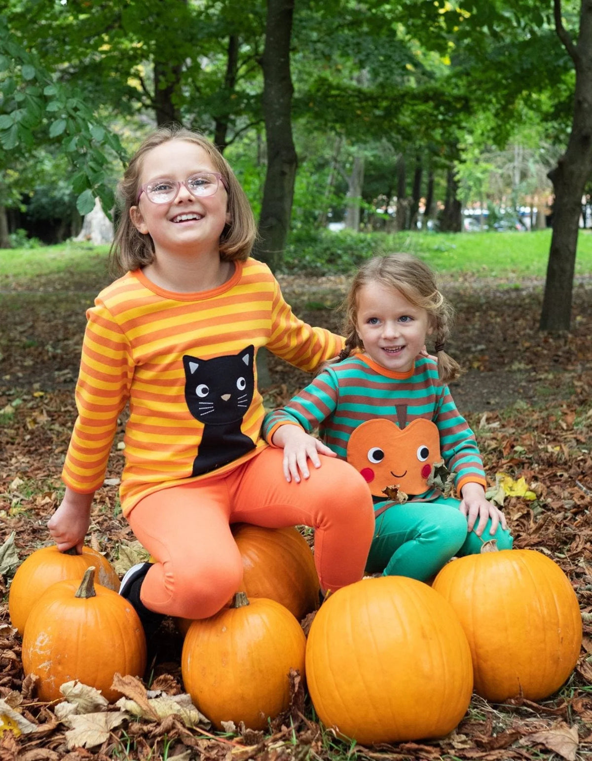 Two children sitting on pumpkins in a park with trees and grass in the background.