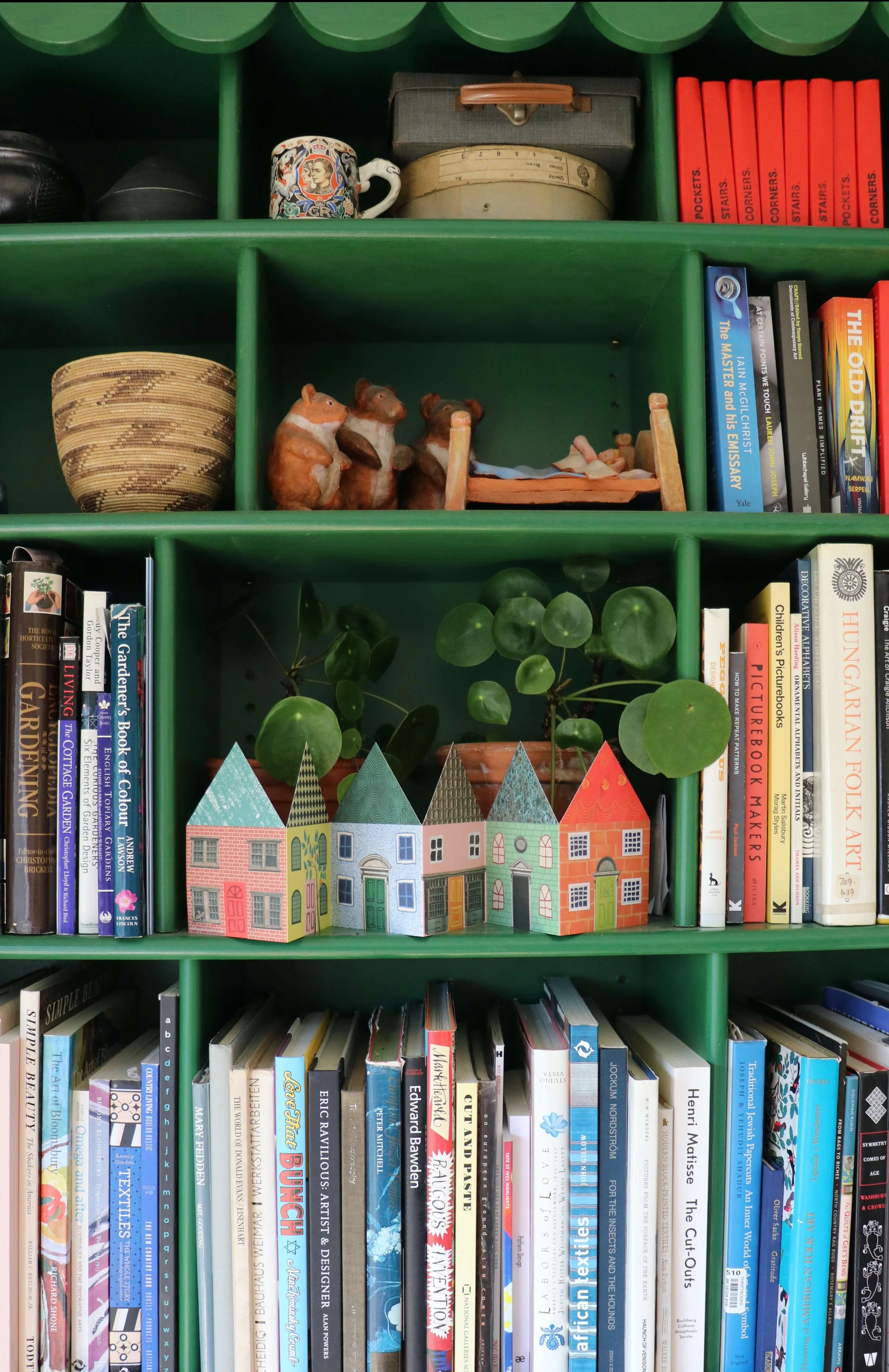Row of miniature paper houses on a green bookshelf.  The houses are colorful and varied in architectural style.