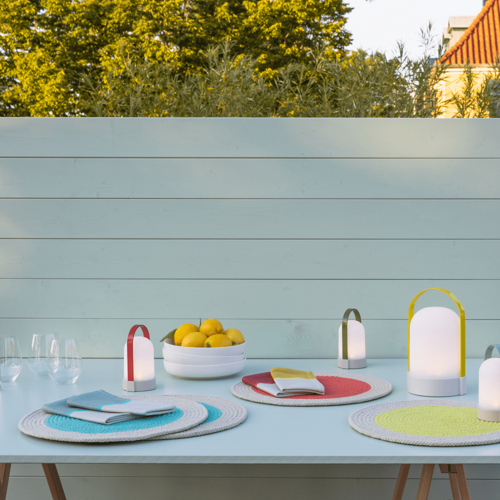 Three portable LED lamps with colored handles on a light blue table, set with placemats and a bowl of lemons.