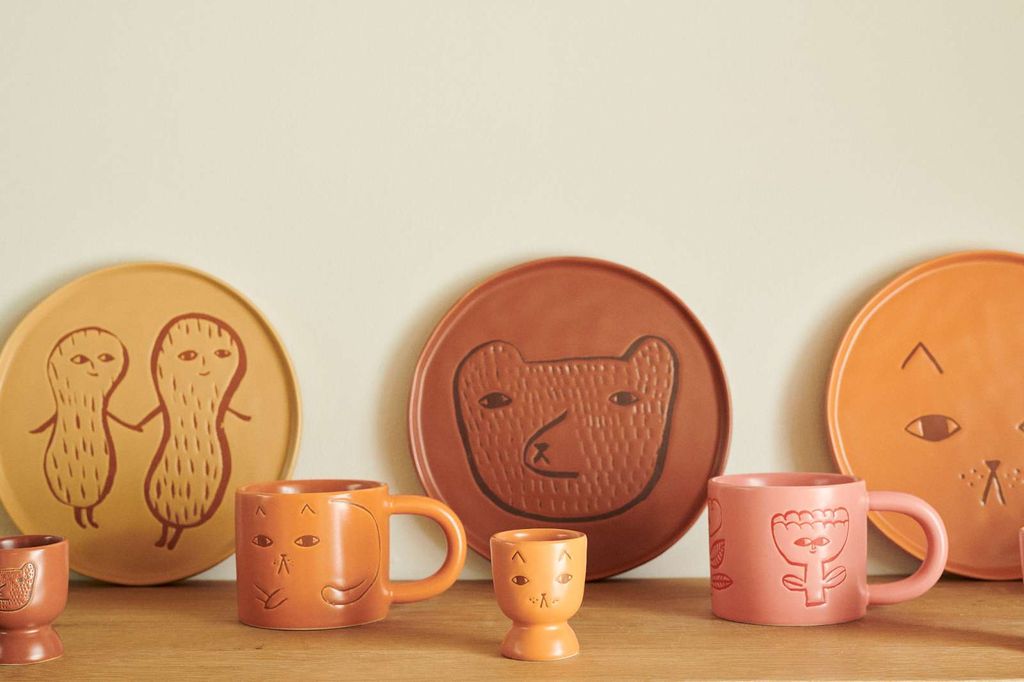 A collection of hand-painted ceramic tableware featuring whimsical animal and plant designs, including orange plates with embossed bear and cat faces, pink and orange mugs, and a small yellow cup, arranged on a wooden surface against a light beige wall.