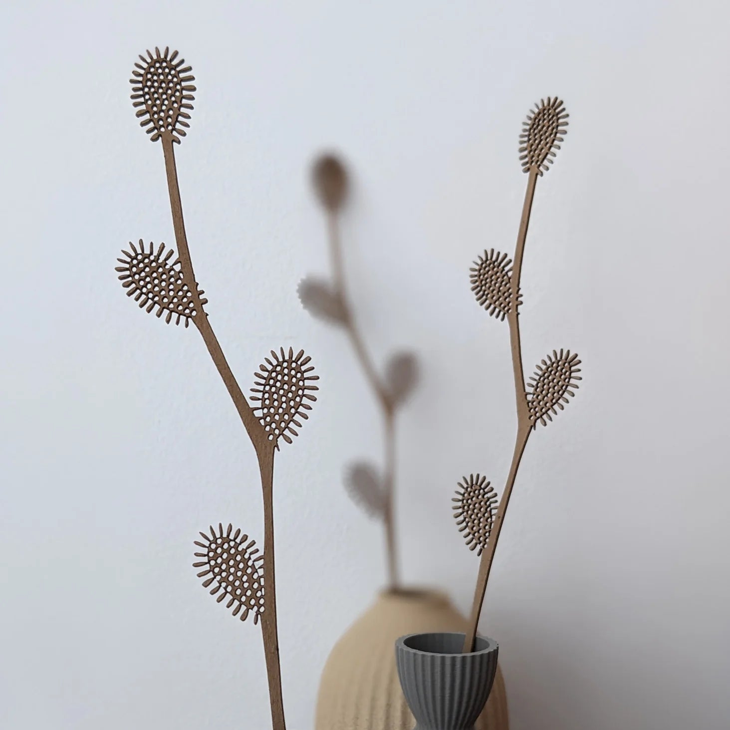Two light brown, laser-cut wood plant stems with textured, spiky leaf details.  The stems are in a gray vase.