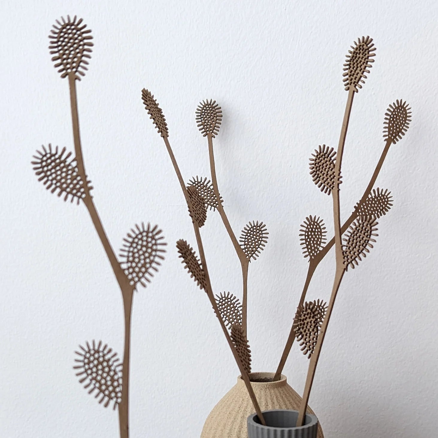 Three brown, laser-cut, decorative plant stems with textured, spherical blossoms in a beige vase.