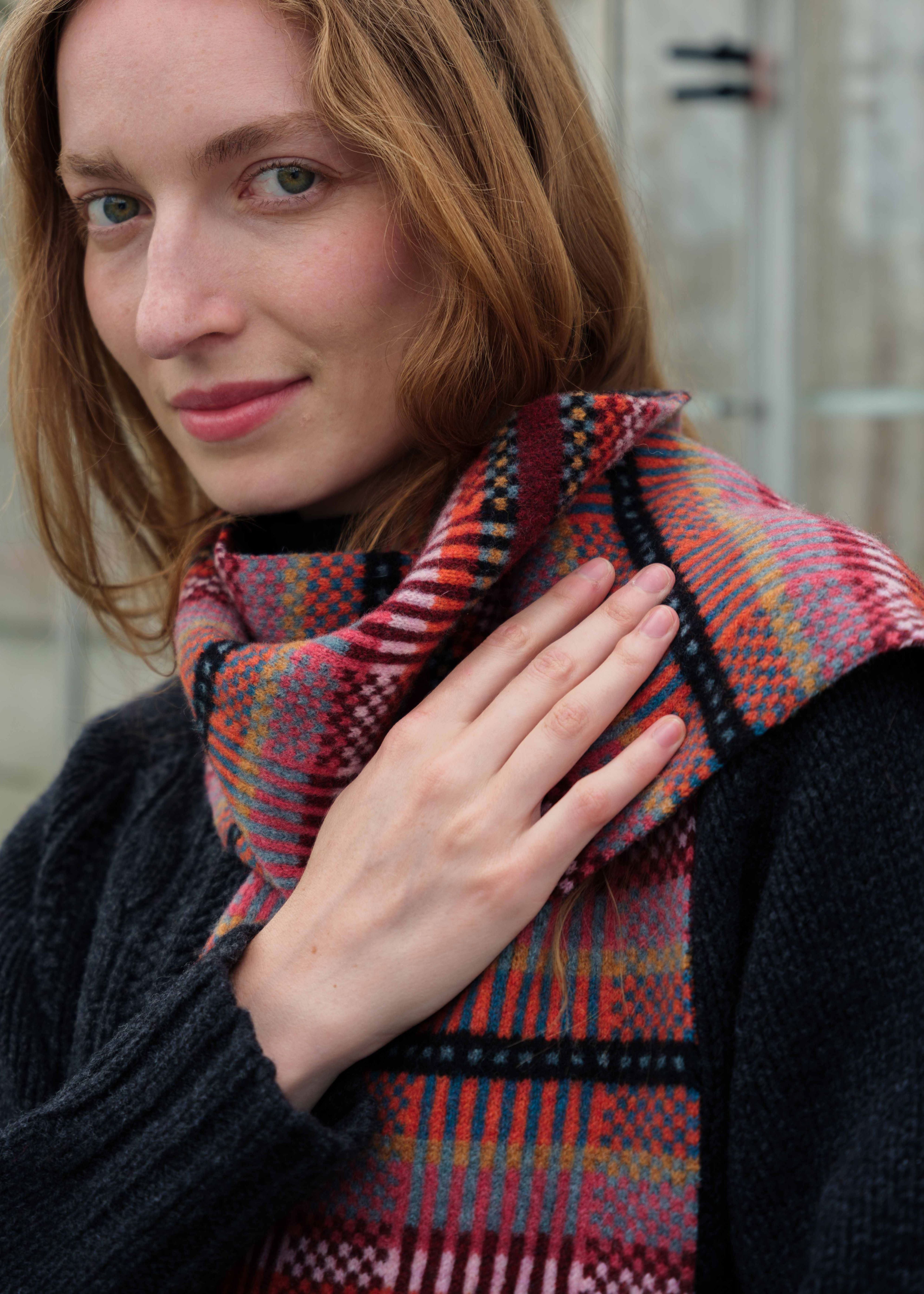 A close-up lifestyle shot of a woman wearing a dark gray knit sweater and a thick, patterned woven scarf in red, orange, black, and yellow.