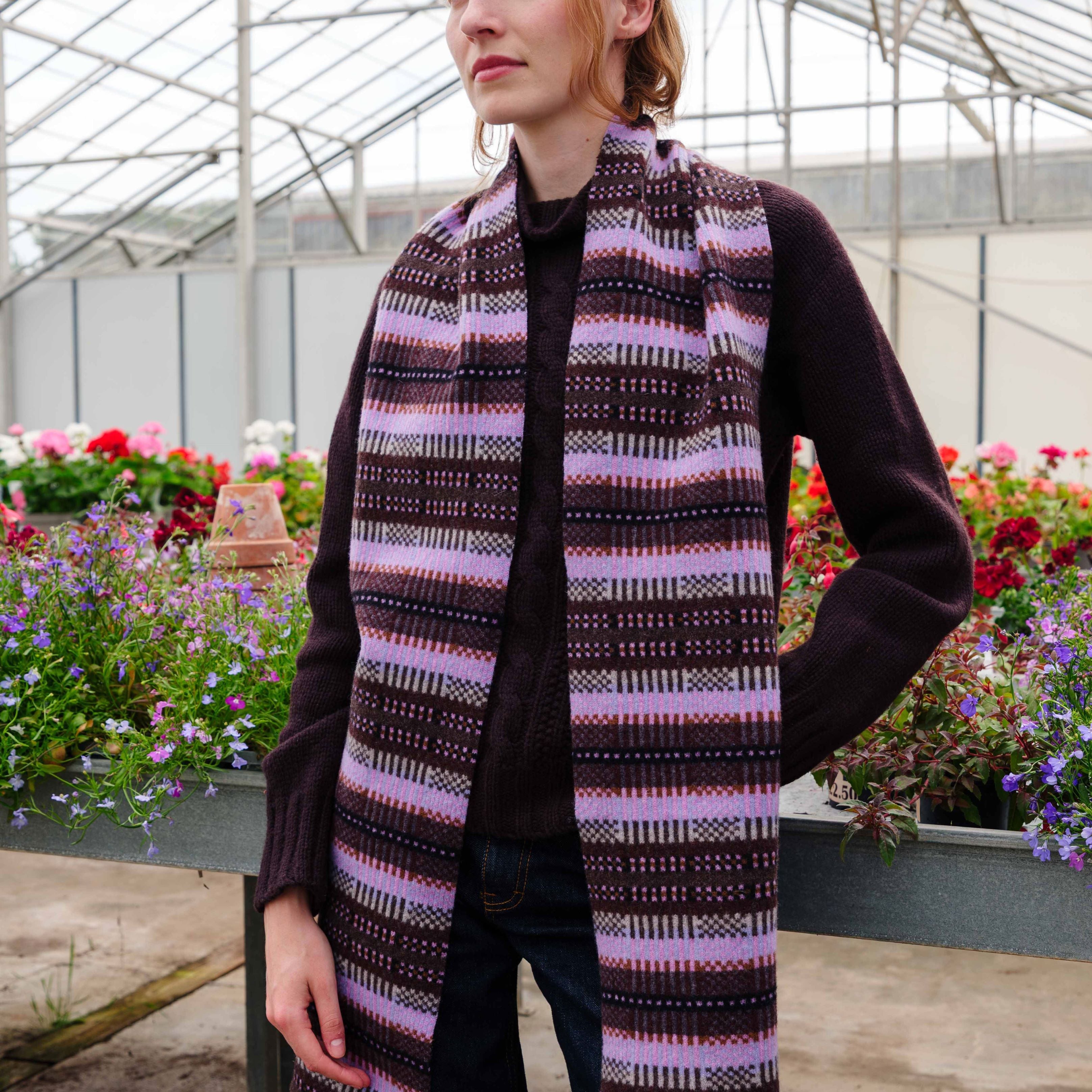 A woman standing inside a greenhouse wearing a long, open-front knit sweater vest with horizontal stripes in purple, pink, and black.