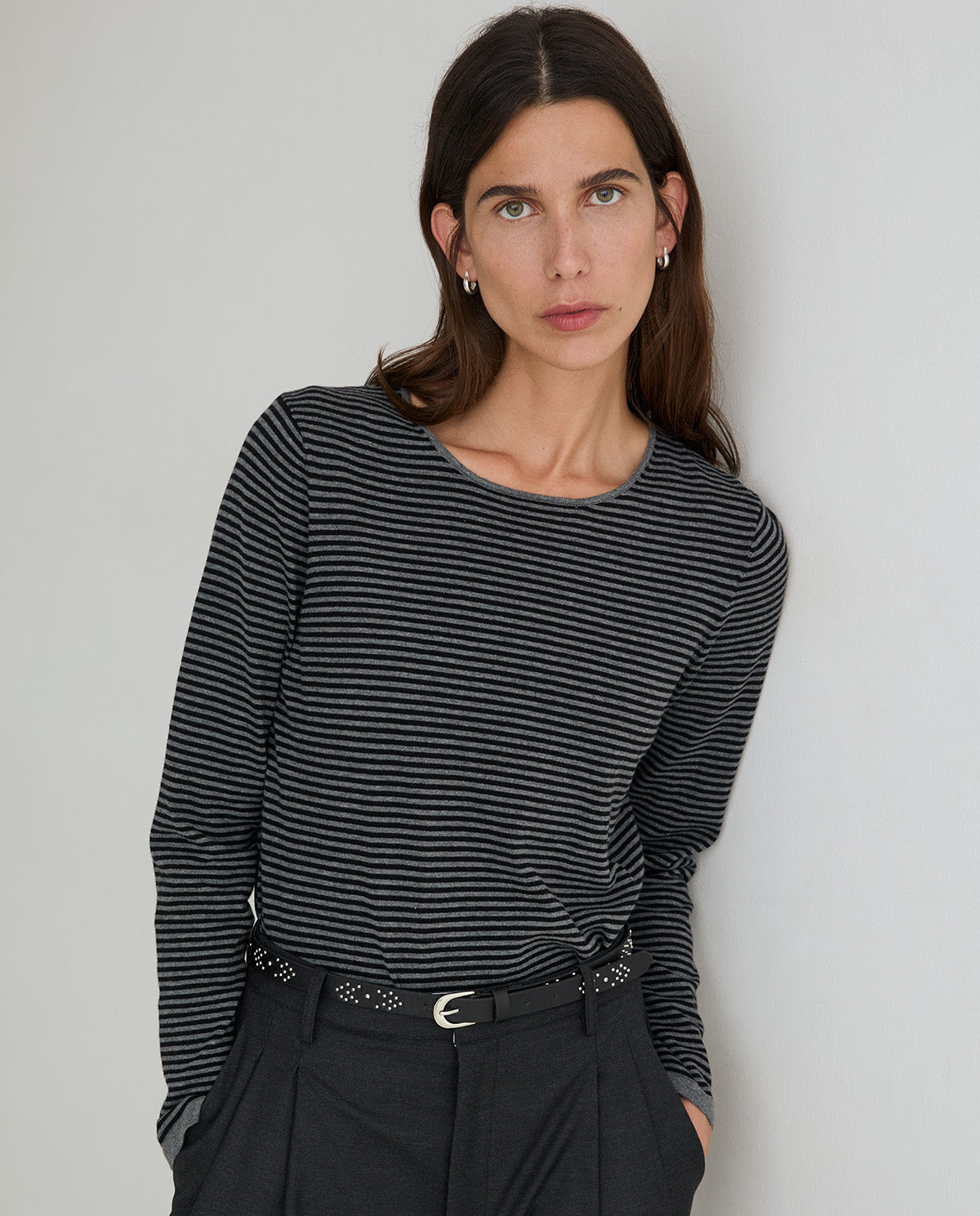 A woman wearing a black and gray striped long-sleeve top with dark trousers, standing against a plain light gray wall.