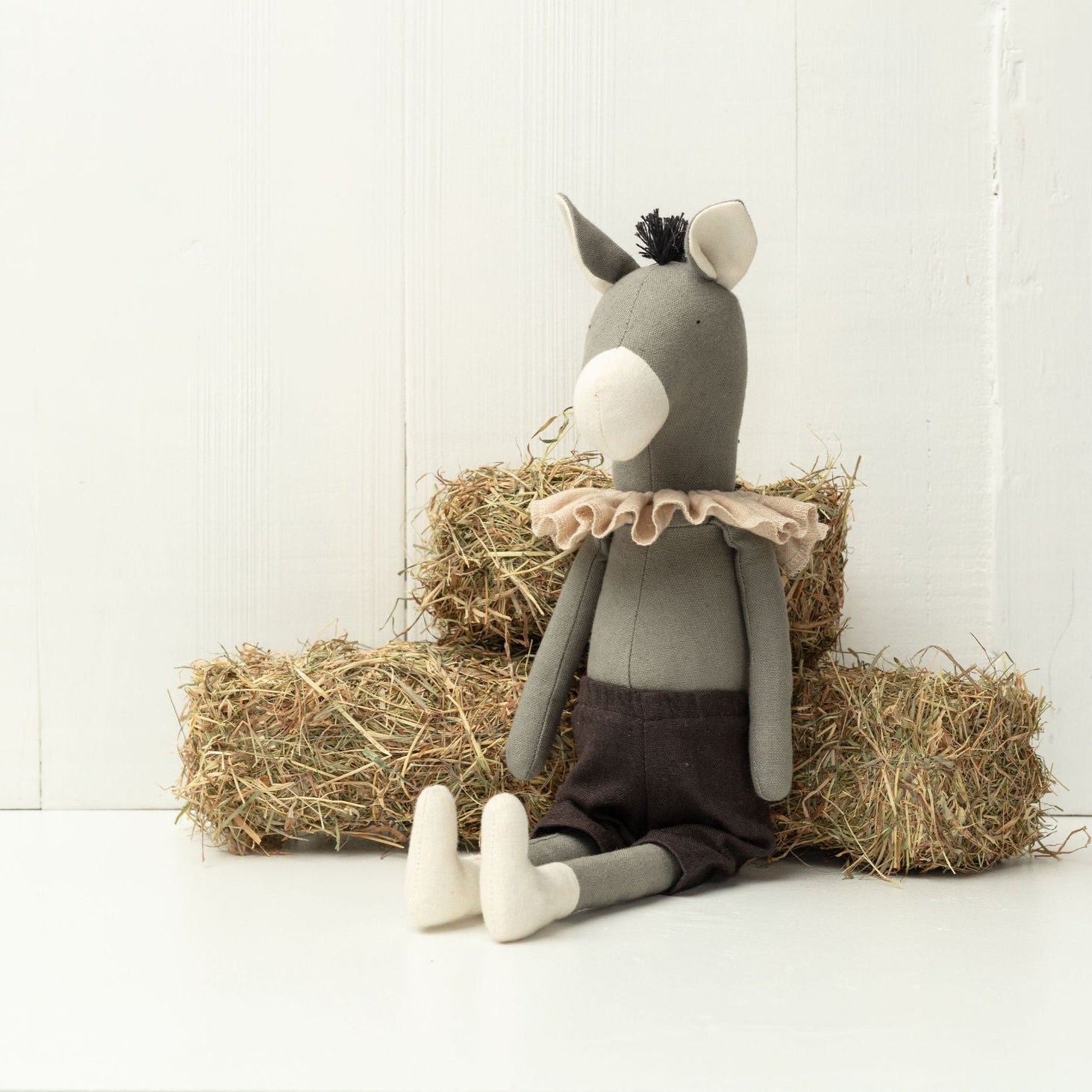 Stuffed donkey toy with a ruffled shirt sitting on hay bales against a white wall.