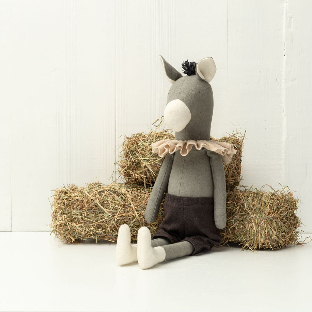 Stuffed donkey toy with a ruffled shirt sitting on hay bales against a white wall.