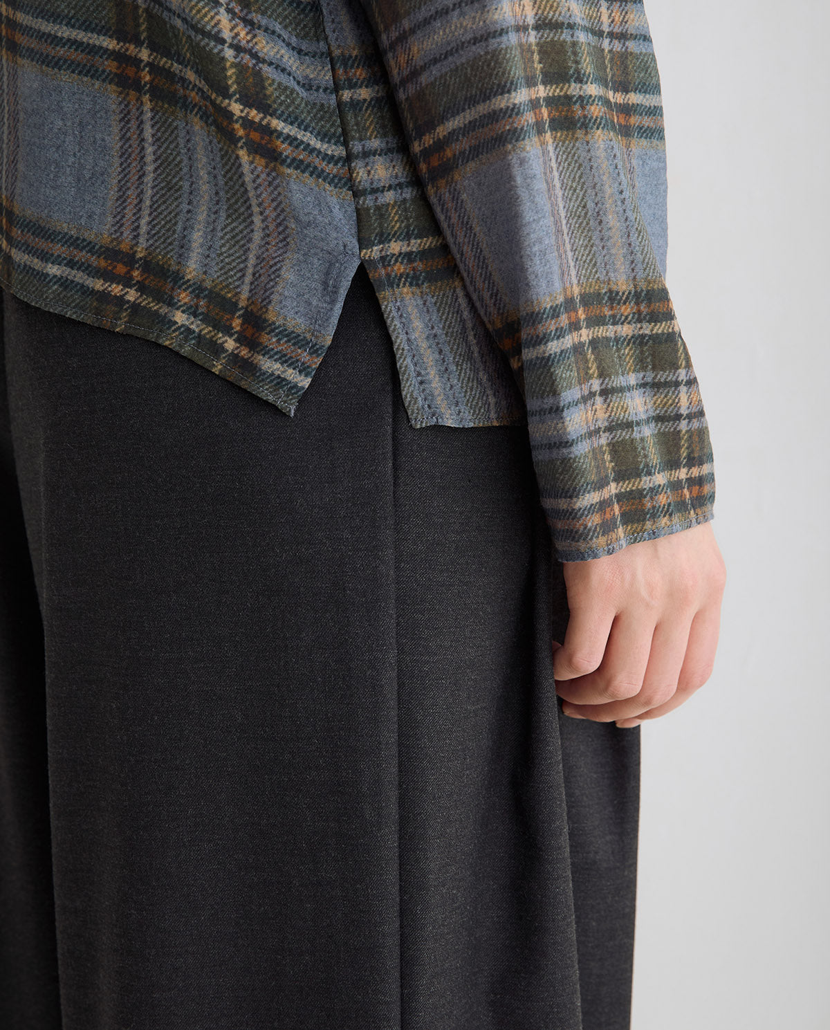 Close-up of a person wearing a muted plaid shirt in blue, olive, and tan tones over a dark charcoal skirt, with one hand visible.