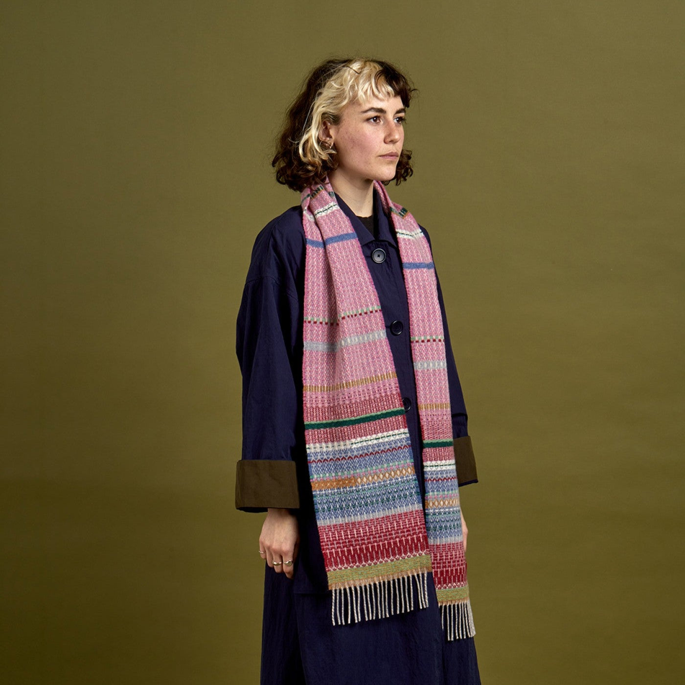 A model wearing a long navy blue coat with brown cuffs, brown wide-leg trousers, and a thick woven scarf in pink, blue, green, and beige, posed against a solid olive green studio background.