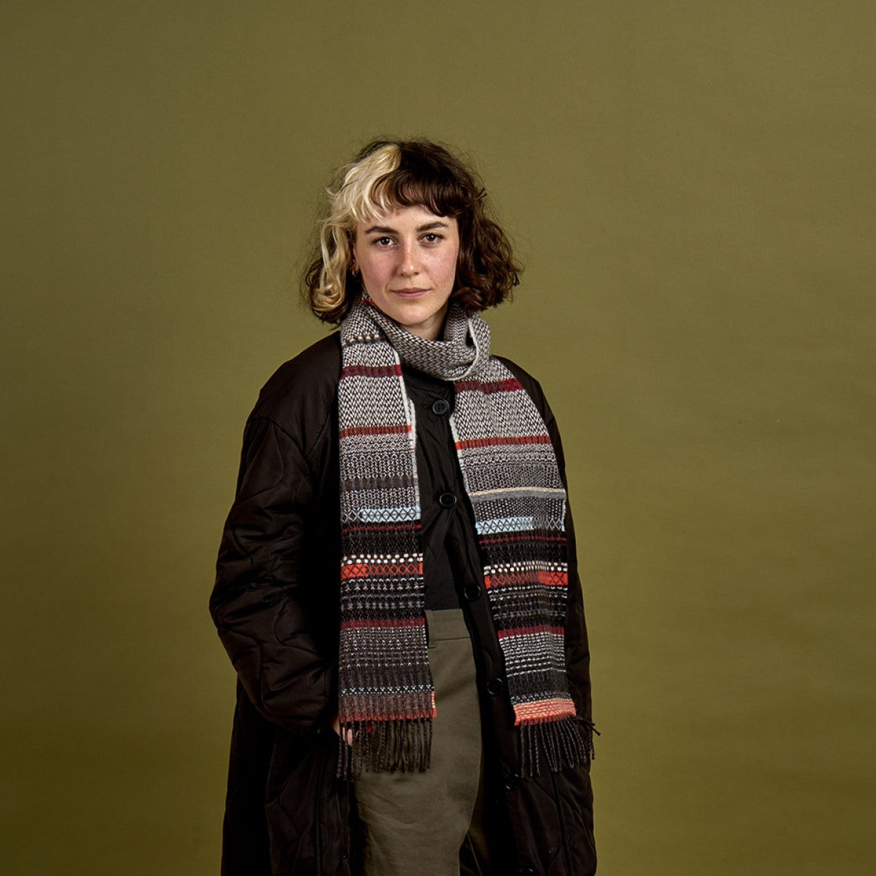 A model stands against a solid olive green studio background wearing a long black quilted coat, a thick gray knit scarf with red, blue, and white geometric stripes, olive green trousers, and black shoes.