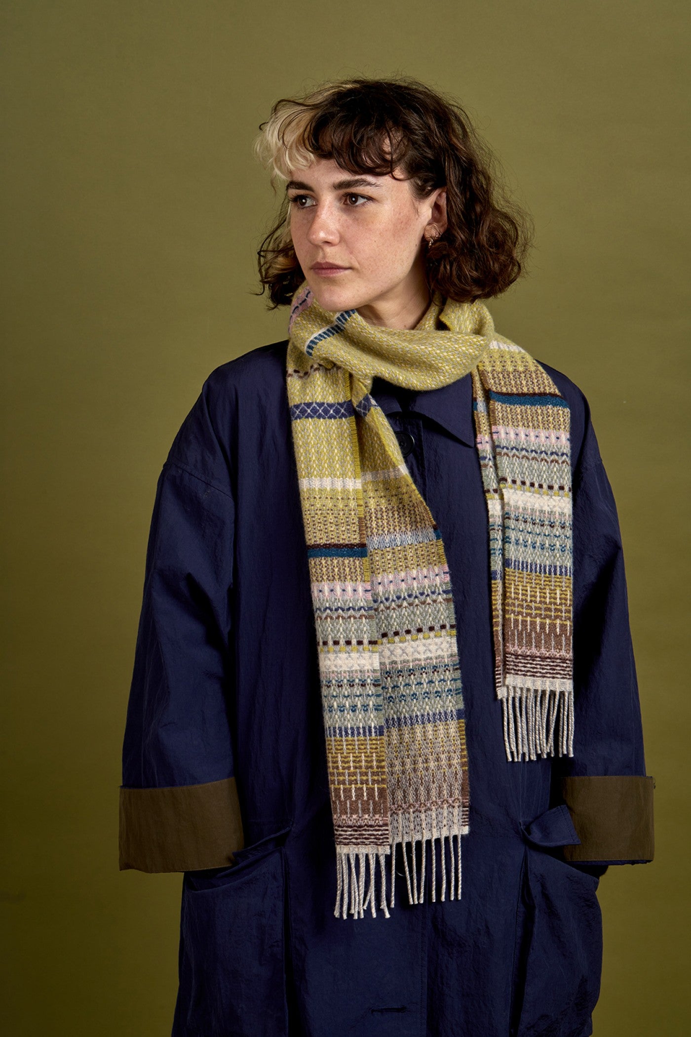 A model wearing a navy blue coat with olive-brown cuff accents and a thick, patterned knit scarf in yellow, blue, and white, set against a solid olive green background.