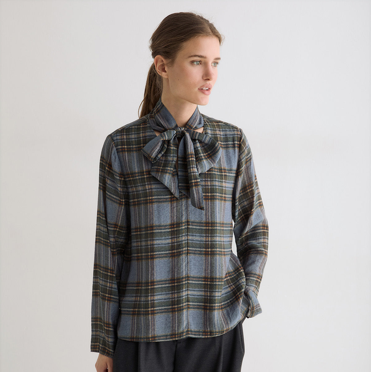 A woman modeling a long-sleeve plaid blouse with a large bow at the collar, paired with dark wide-leg trousers, against a plain light gray studio background.