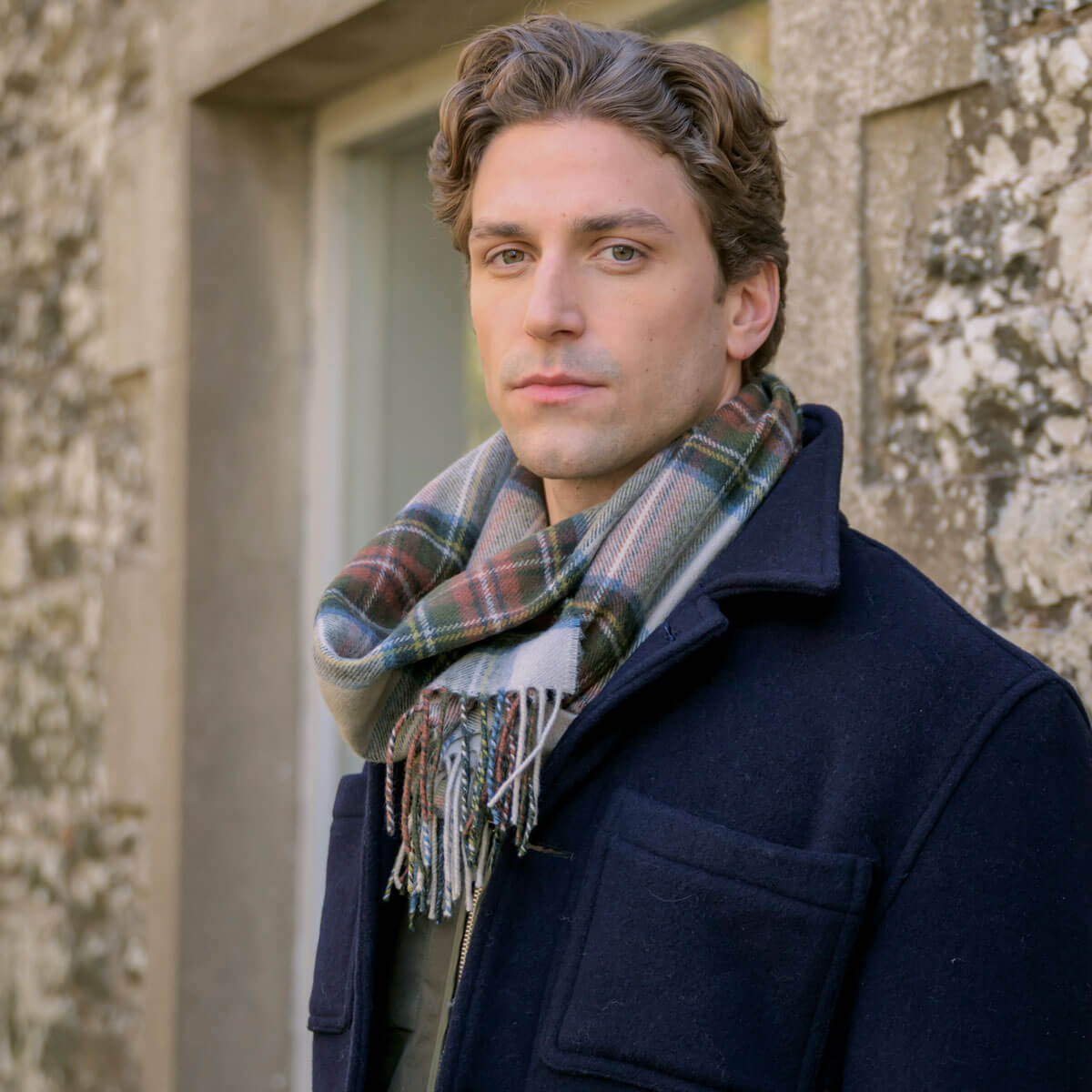 Man wearing a dark coat and plaid scarf standing against a stone wall.