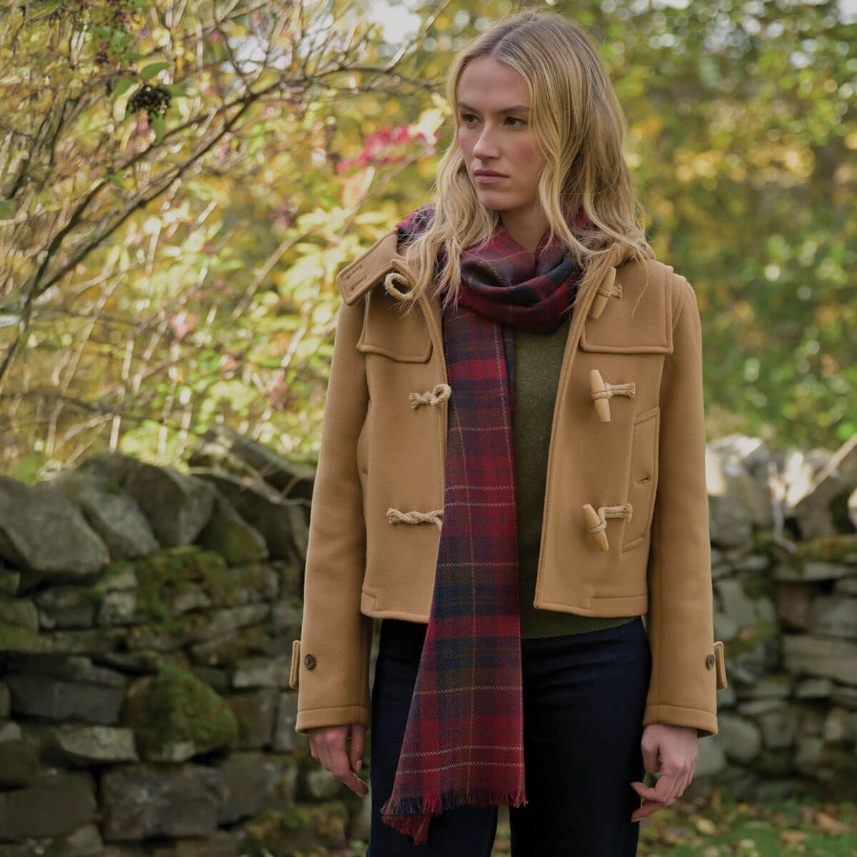 Woman wearing a tan coat and red plaid scarf standing in a natural setting with stone walls and trees.