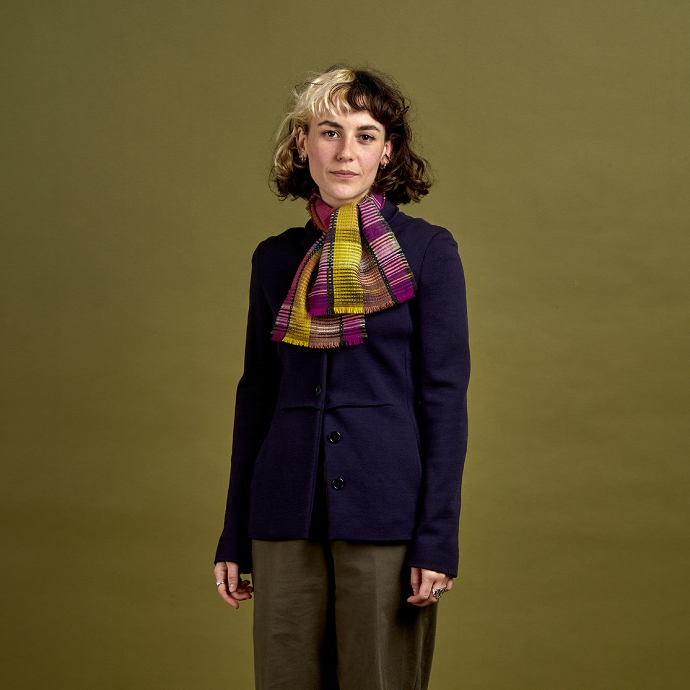 A woman stands against a solid olive green studio background wearing a dark navy blazer, olive brown wide-leg trousers, and a vibrant striped scarf in yellow, purple, and pink.
