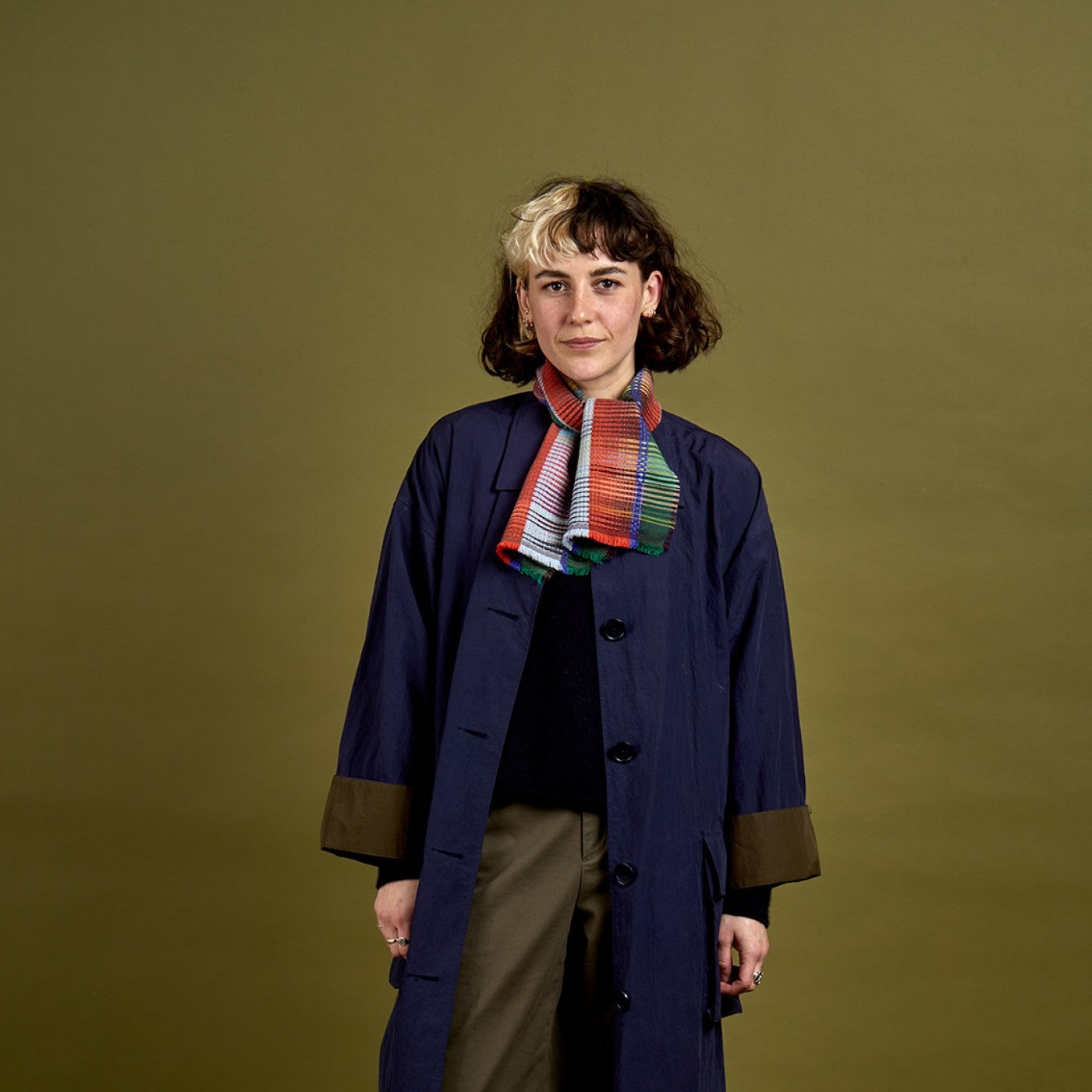A model stands against a solid olive green studio background wearing a long navy blue coat with brown cuffs, brown trousers, a colorful striped knit scarf, and black shoes.