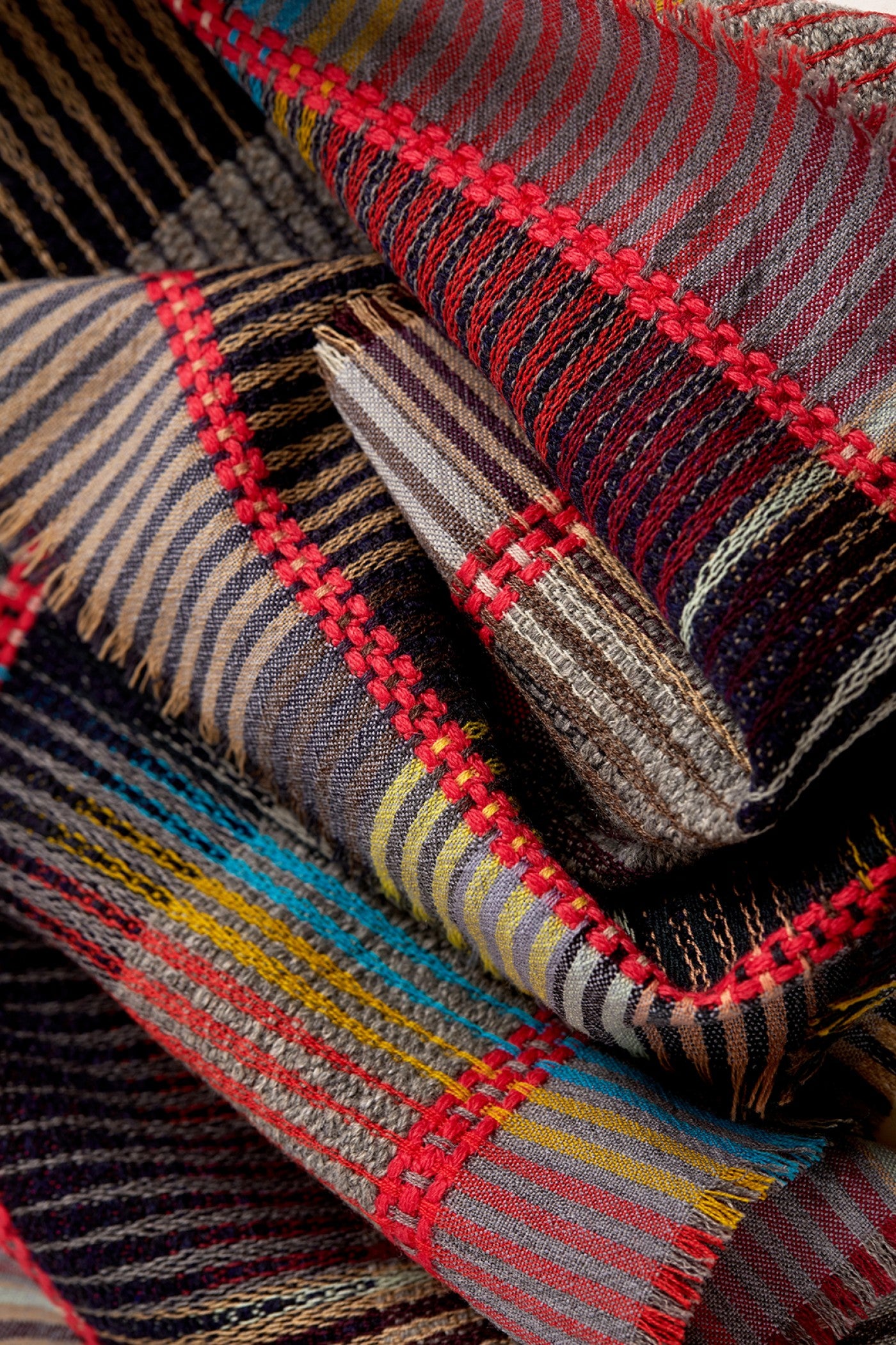 A close-up of a folded plaid woven textile, likely a scarf or throw blanket, showing a multicolor pattern with red, blue, yellow, brown, and black stripes.