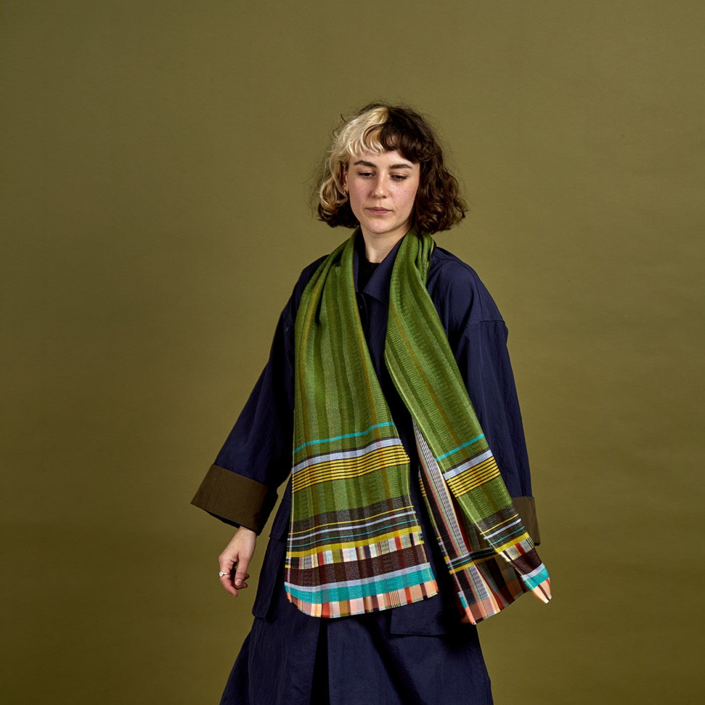Person wearing a long dark blue coat and a green scarf with a plaid pattern on an olive green background