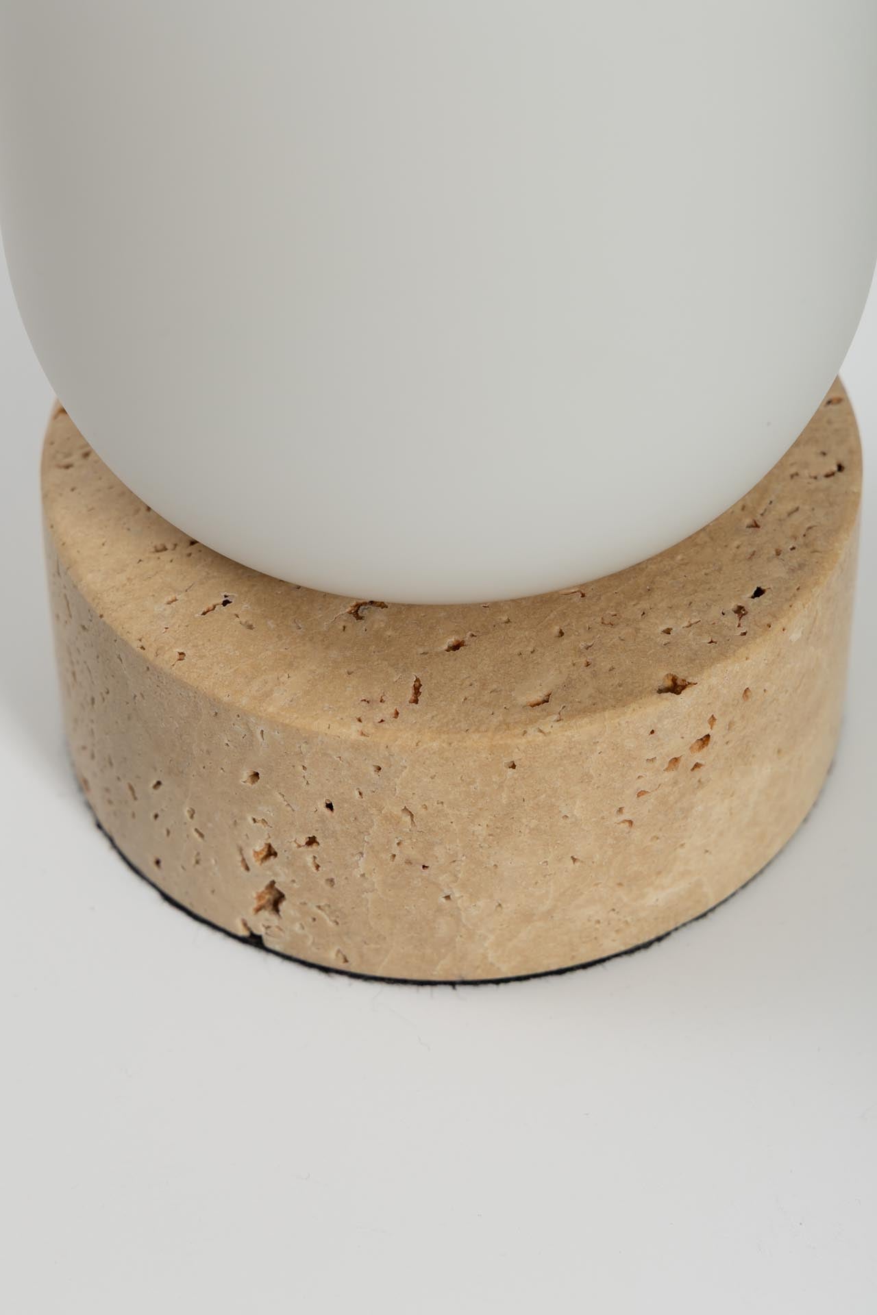 Close-up of a modern table lamp base featuring a round, textured beige travertine stone pedestal supporting a matte white ceramic pot.