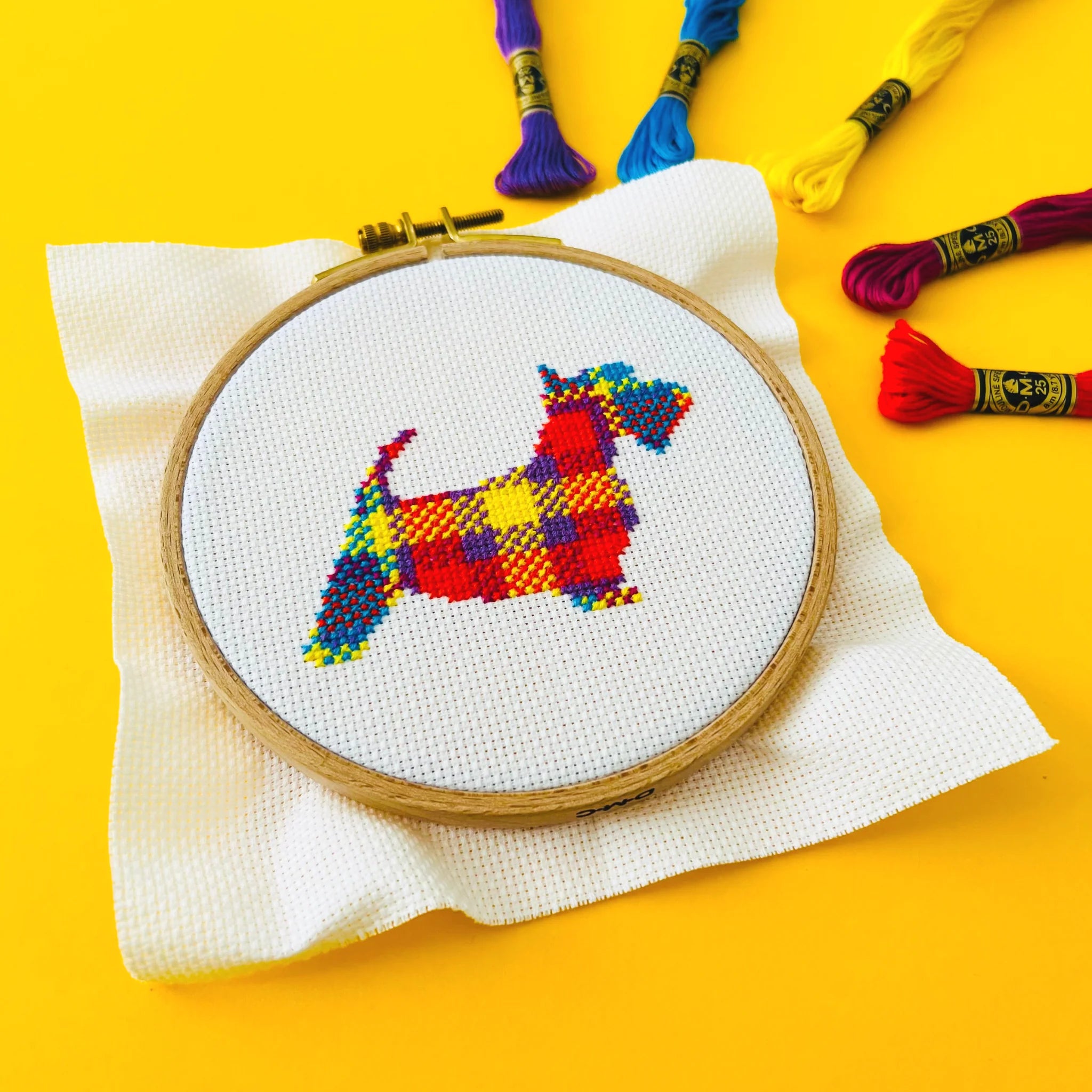 Cross-stitch embroidery in a wooden hoop depicting a colorful, plaid Scottish Terrier.