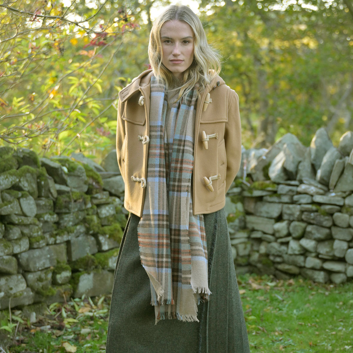 Woman wearing a beige coat and plaid scarf standing in front of a stone wall.