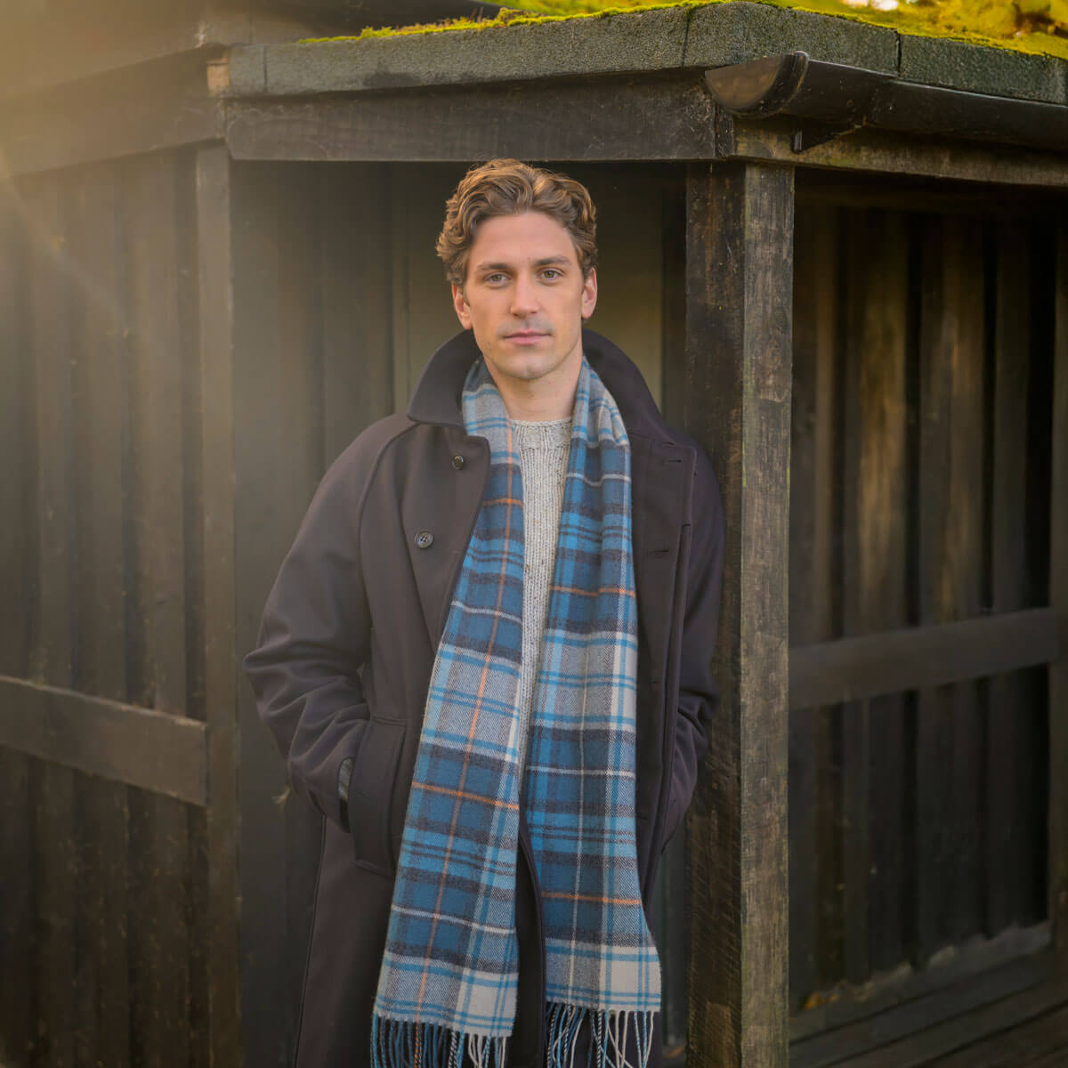 Man wearing a dark coat and blue plaid scarf standing in front of a wooden structure.
