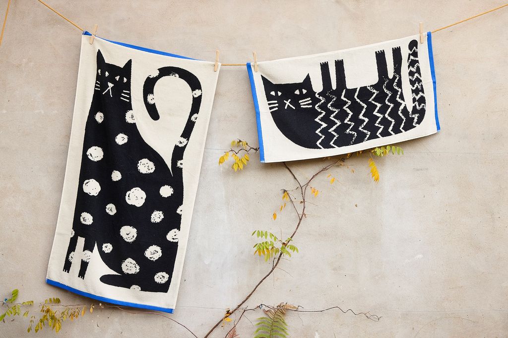 Two decorative towels with cat designs hanging on a line against a neutral background.