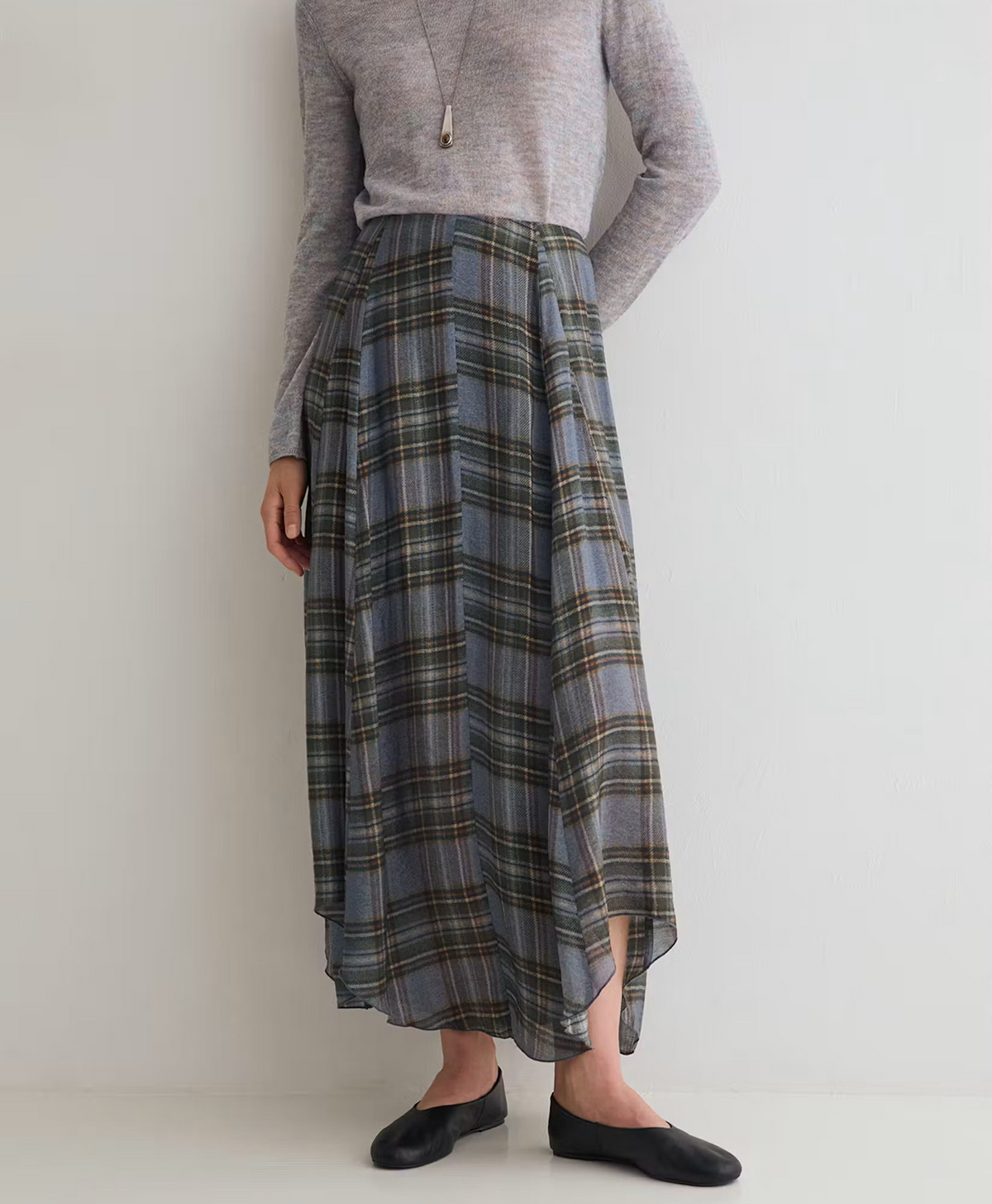 A model wearing a gray long-sleeve knit top and a high-waisted, pleated plaid midi skirt in shades of gray, blue, and green, paired with black flats.