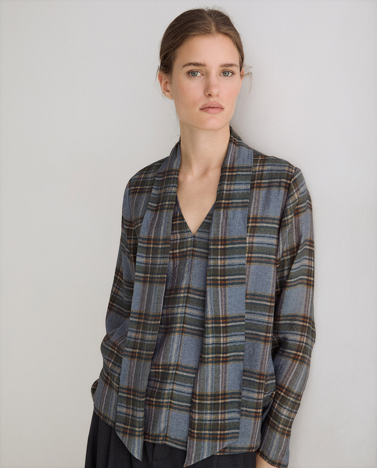 A woman wearing a blue-gray plaid wrap-style blouse with brown and beige stripes, photographed against a plain light gray studio background.