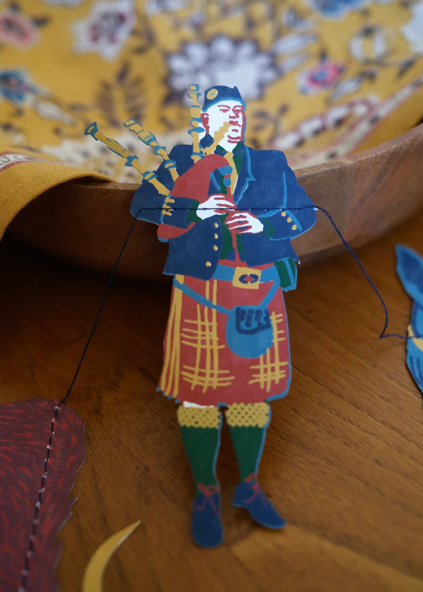 Hand-stitched paper cutout of a bagpiper in a kilt, playing bagpipes.  The figure is colorful and detailed.