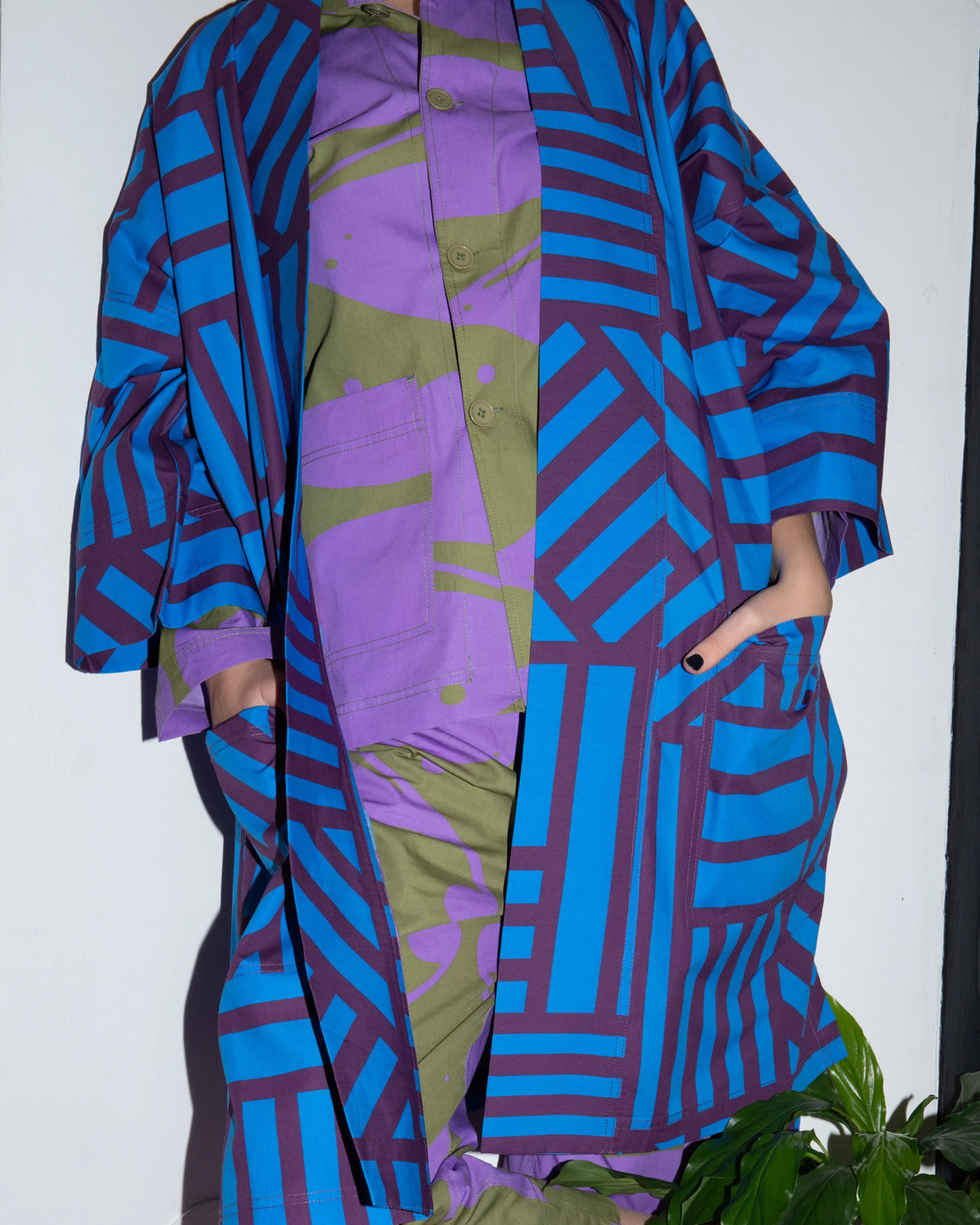 A person wearing a colorful patterned outfit consisting of a blue and purple geometric open-front jacket over a purple and olive green abstract-print shirt and pants, standing against a plain white wall.
