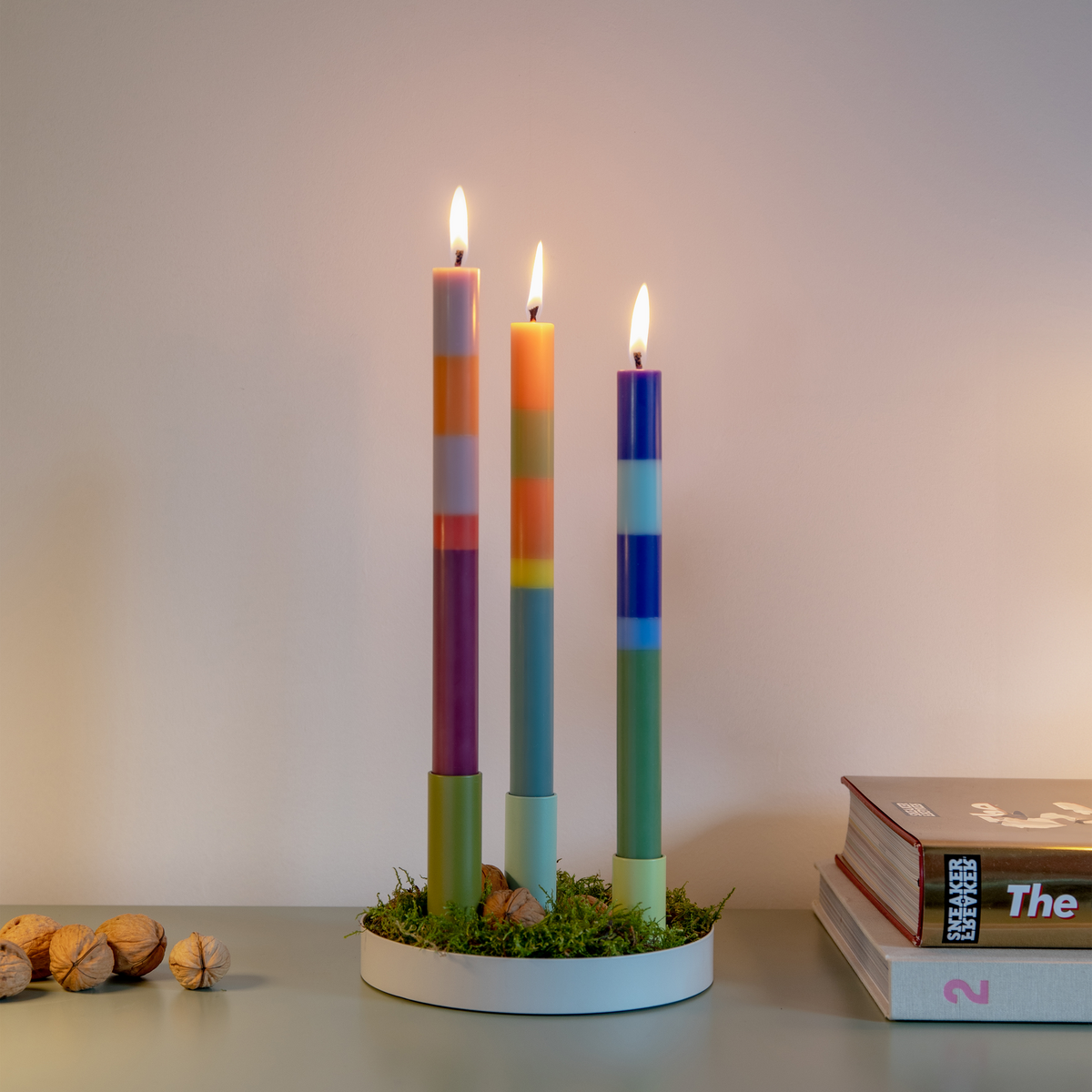 Three colorful candles in a holder on a table with books and nuts.