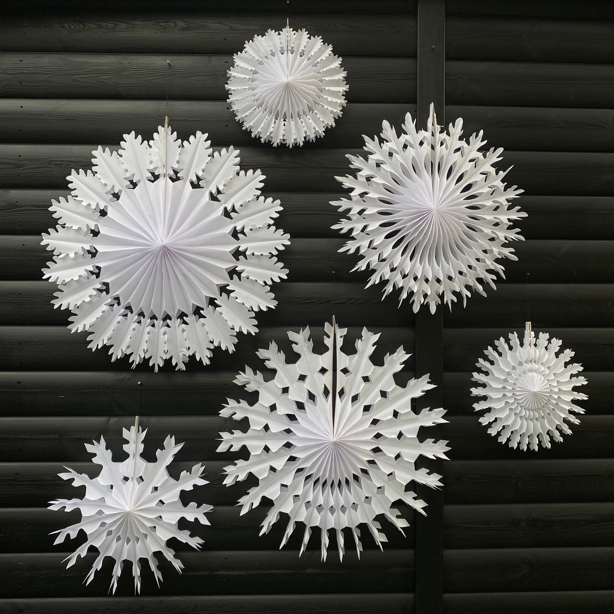 Five large and two small white paper snowflake decorations against a dark wood background.