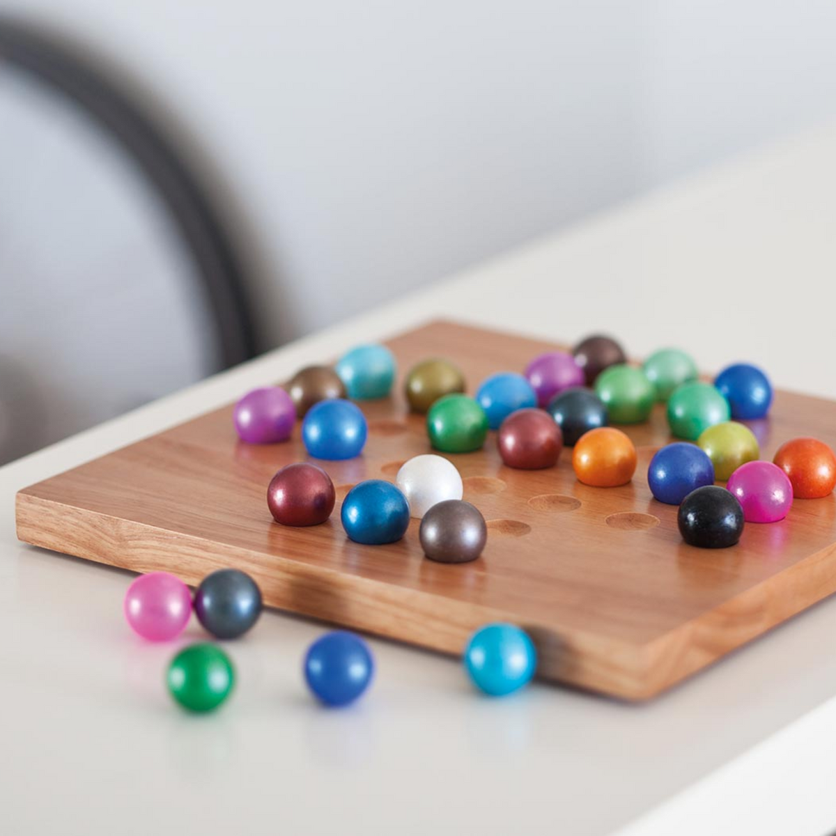 A wooden square board with colorful glossy round balls scattered on and around it, photographed on a light tabletop with a softly blurred background.