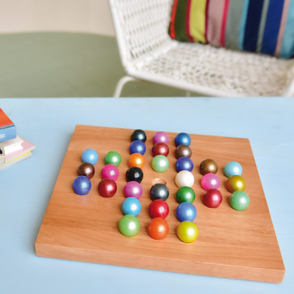 A wooden board filled with colorful glossy round beads, photographed on a light blue tabletop with a blurred white chair and striped pillow in the background.