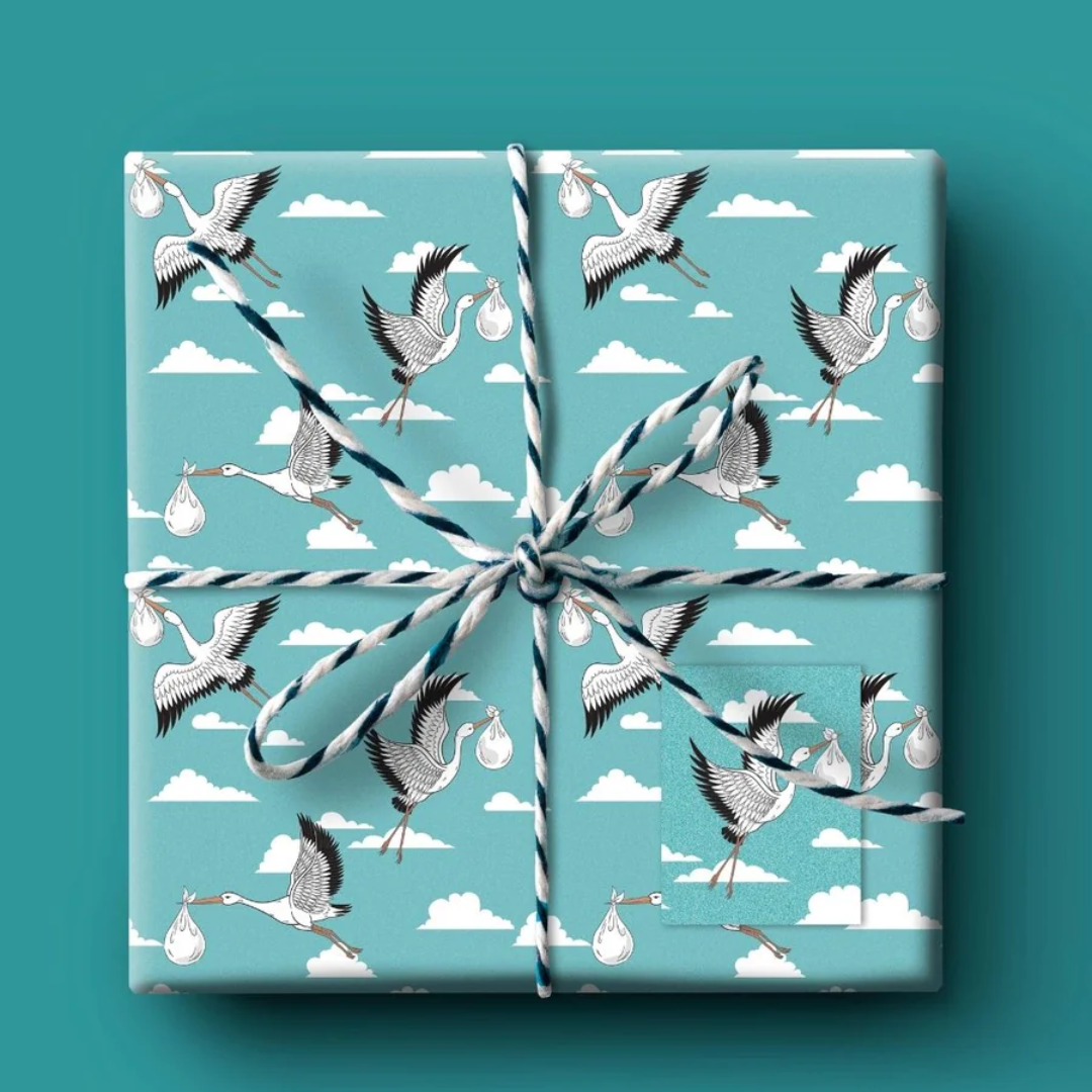 Gift wrap featuring a repeating pattern of storks carrying bundles against a light blue sky with white clouds.