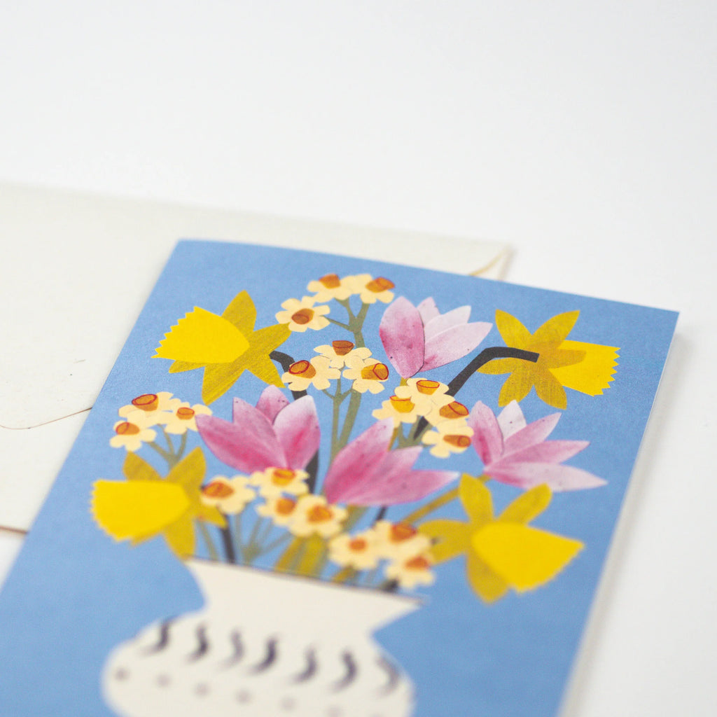 A greeting card featuring a stylized illustration of yellow daffodils and pink tulips in a white vase against a light blue