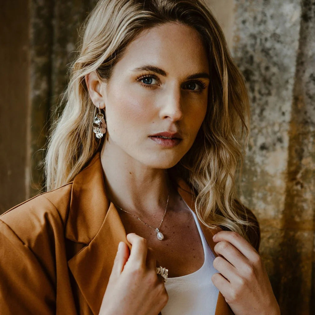 Woman wearing a delicate pearl necklace and layered disc earrings.  She is also wearing a brown blazer.