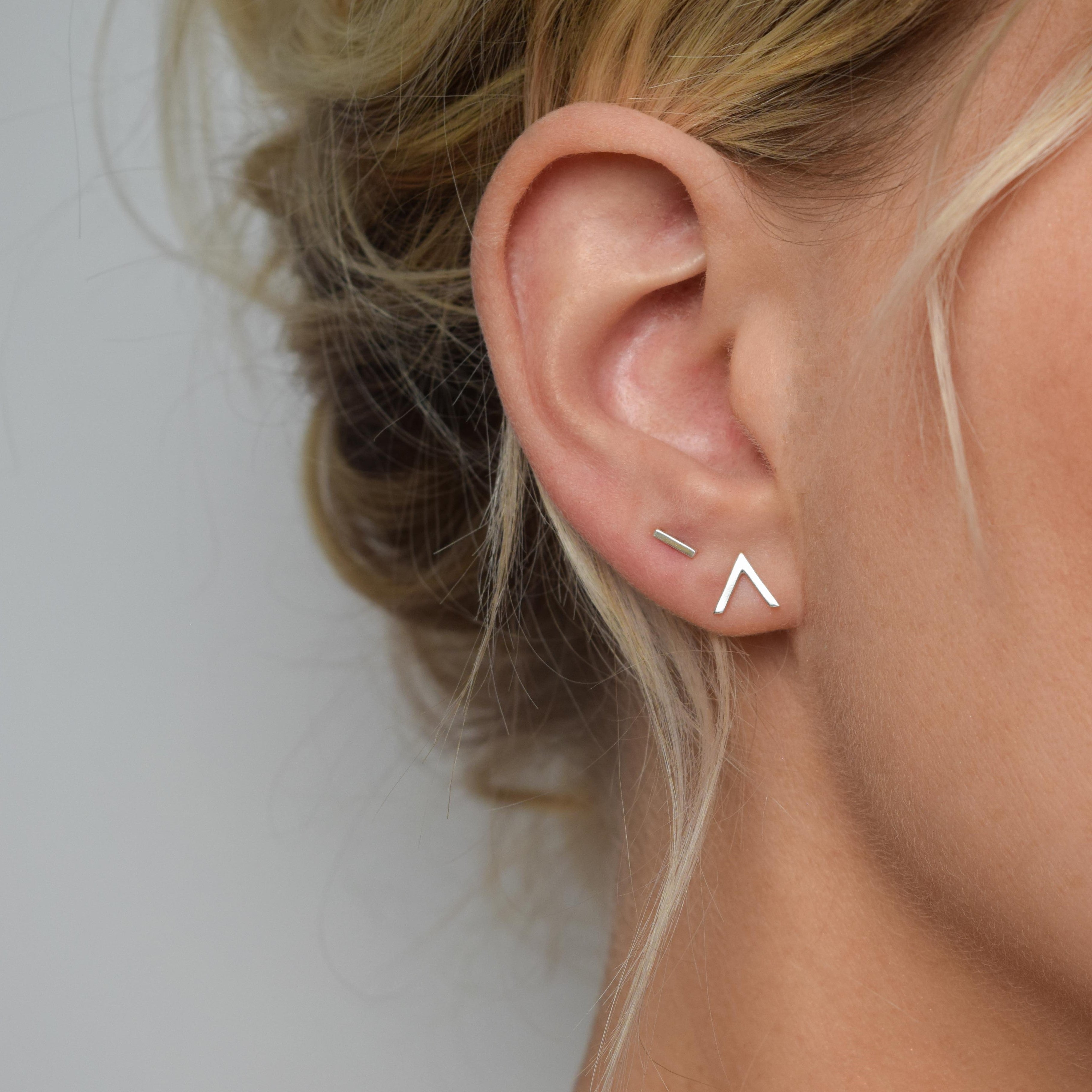 Close-up of a woman's ear wearing a pair of minimalist silver stud earrings; one is a short bar, the other a small inverted