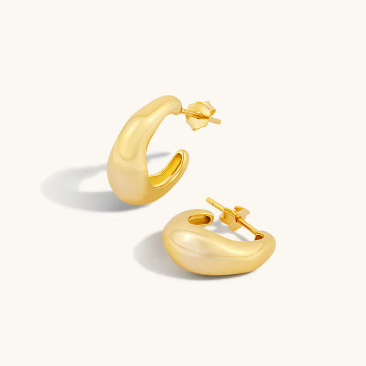A pair of polished gold huggie hoop earrings with a rounded, organic shape and post-back closures, displayed on a clean off-white background.