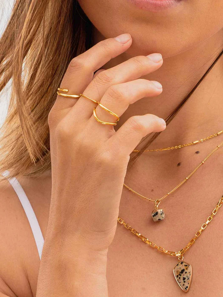 Close-up of a model wearing layered gold necklaces and rings, including a teardrop-shaped stone pendant necklace and a double-ring stack, with her hand near her face.