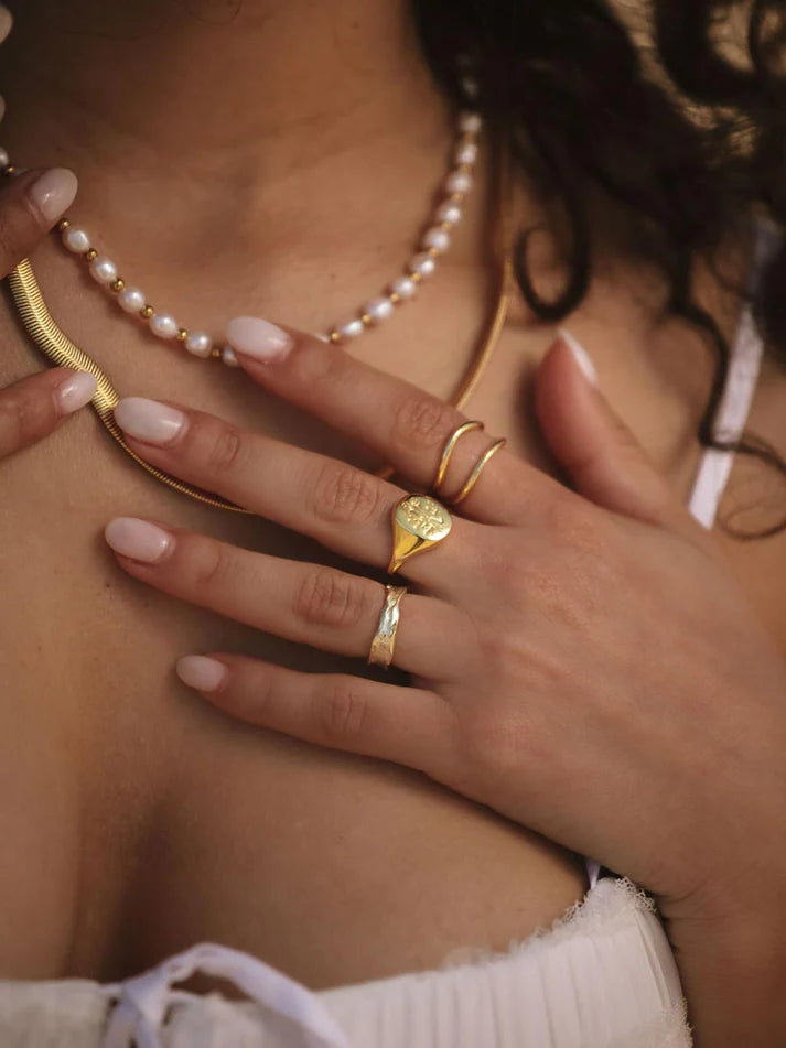 A close-up lifestyle shot of a model wearing multiple gold rings, including a signet-style ring with a round face, a thin wrap-around band, and a textured band, along with a pearl necklace.
