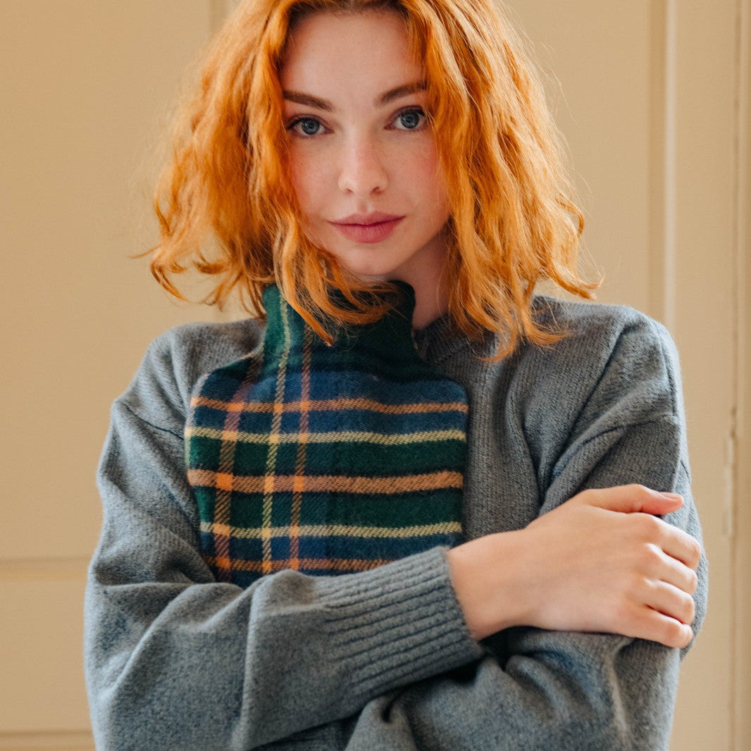 Woman wearing a gray sweater over a plaid shirt with red hair, standing against a beige wall.
