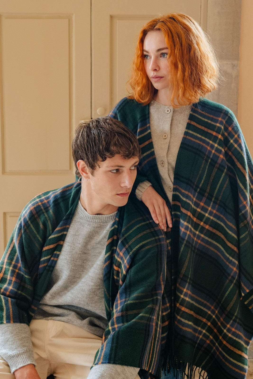 Two people sitting together under a plaid blanket in a warm, indoor setting.