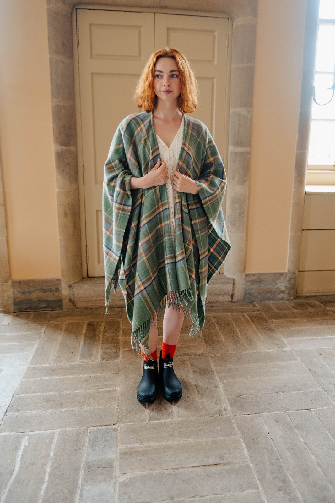 Woman wearing a green plaid poncho in a stone-walled room.