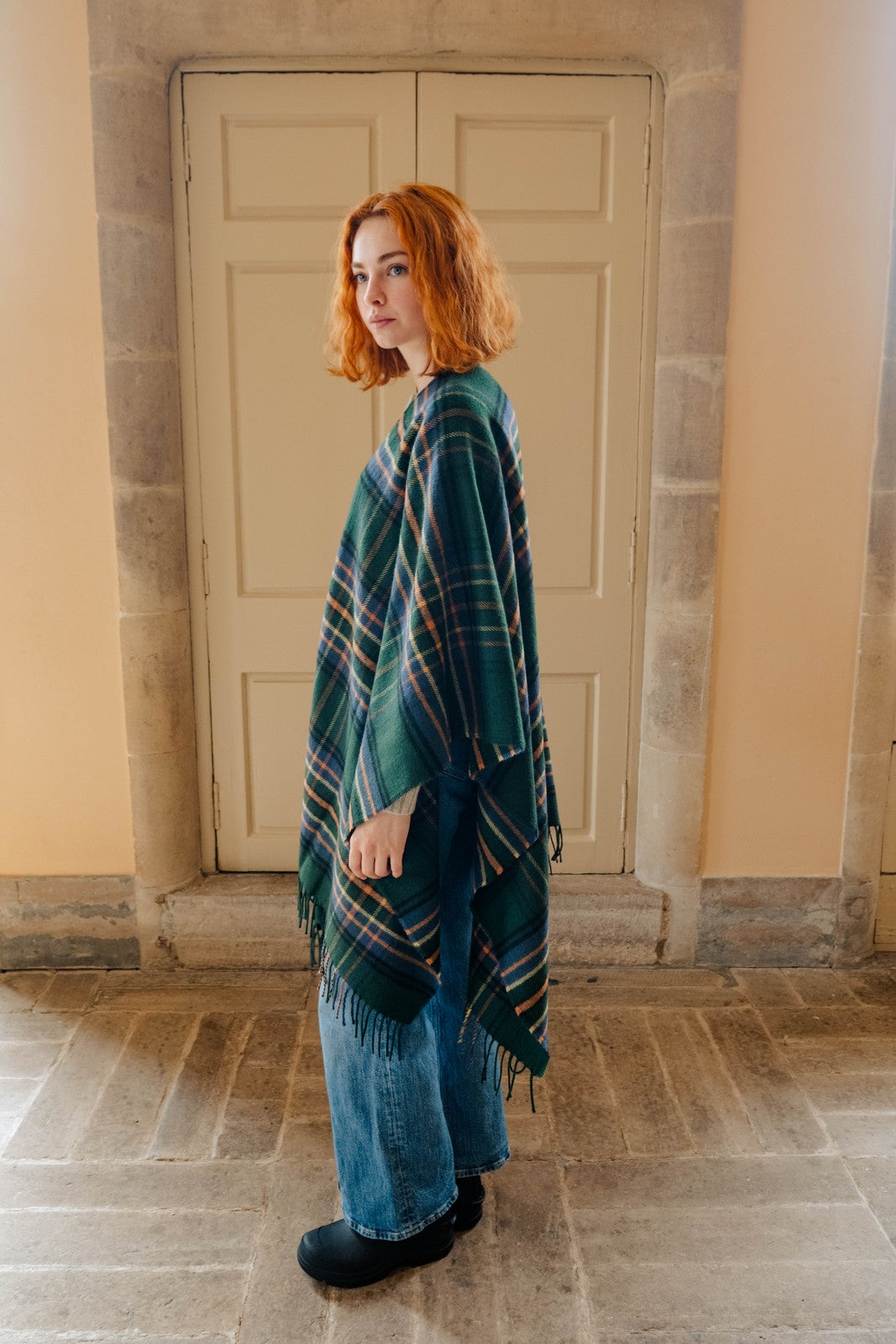 Woman wearing a green plaid shawl standing in front of a stone wall.