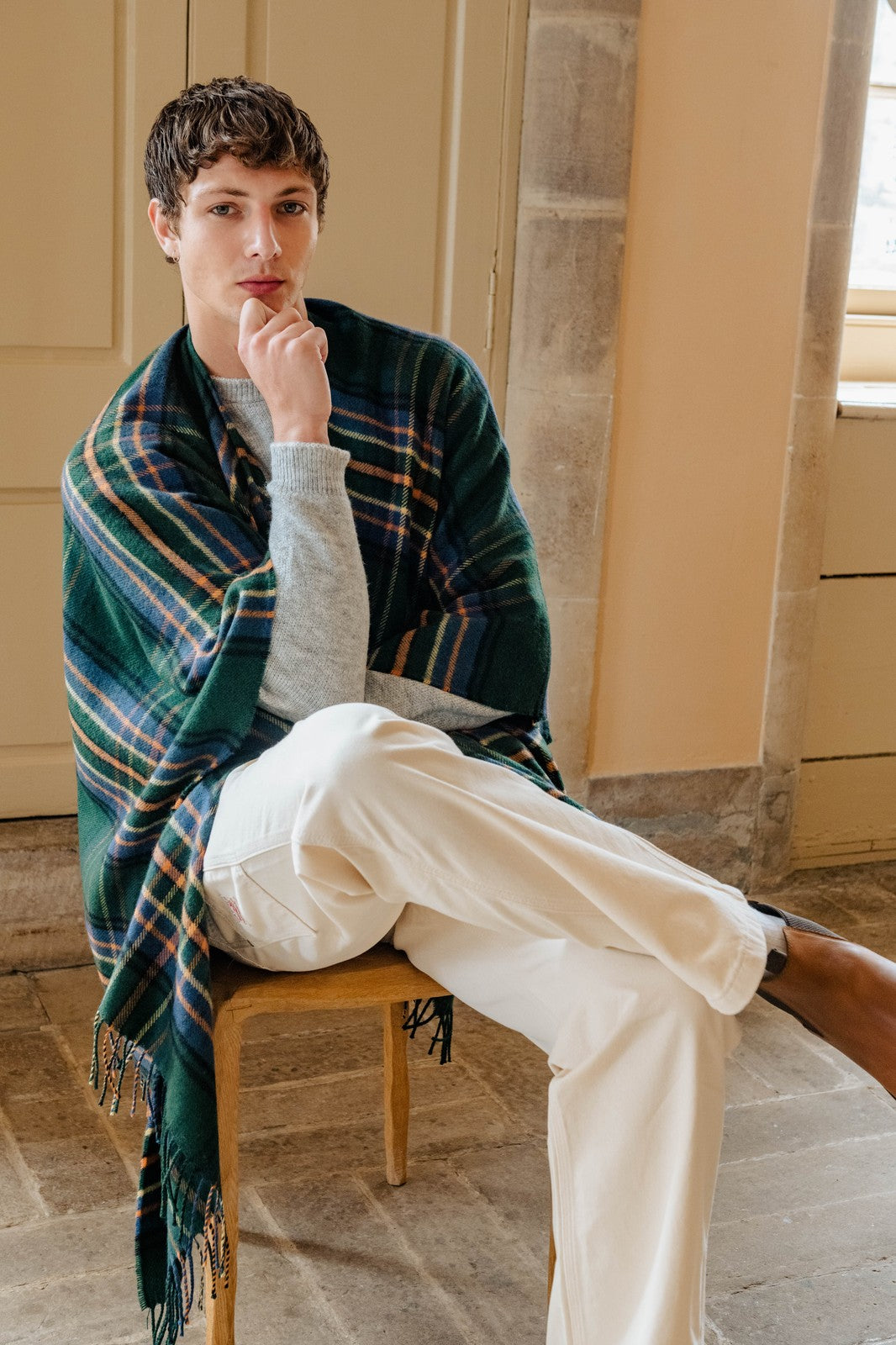 Man sitting on a chair wearing a plaid blanket, gray sweater, and white pants in a room with beige walls.