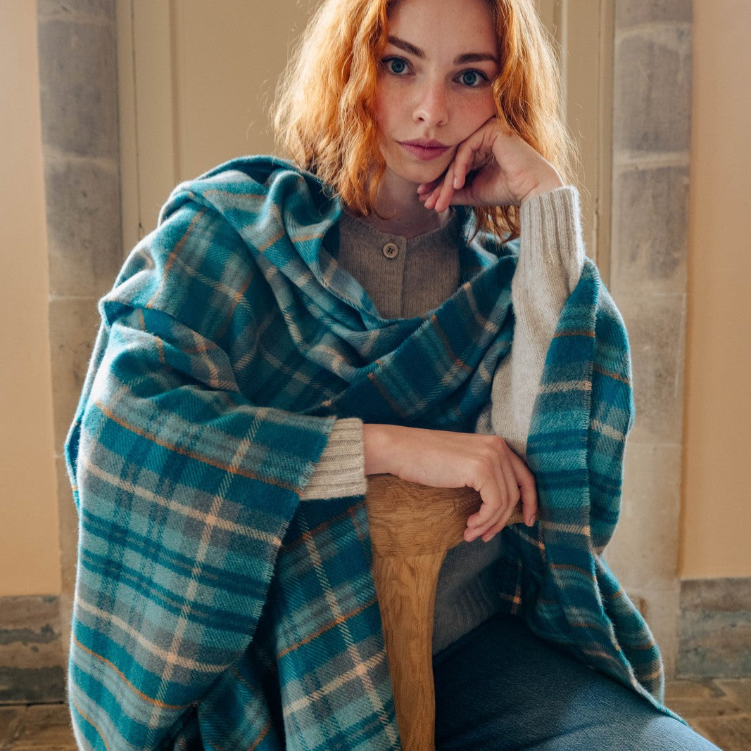 Woman sitting on a wooden stool wearing a blue plaid blanket draped over her shoulders.